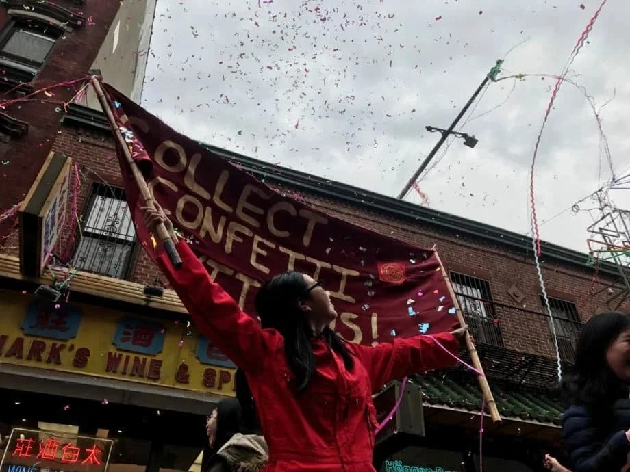 A woman in a red jacket holding a large burgundy banner with white text during a street protest. The crowd is surrounded by confetti and streamers, with buildings and a cloudy sky in the background.