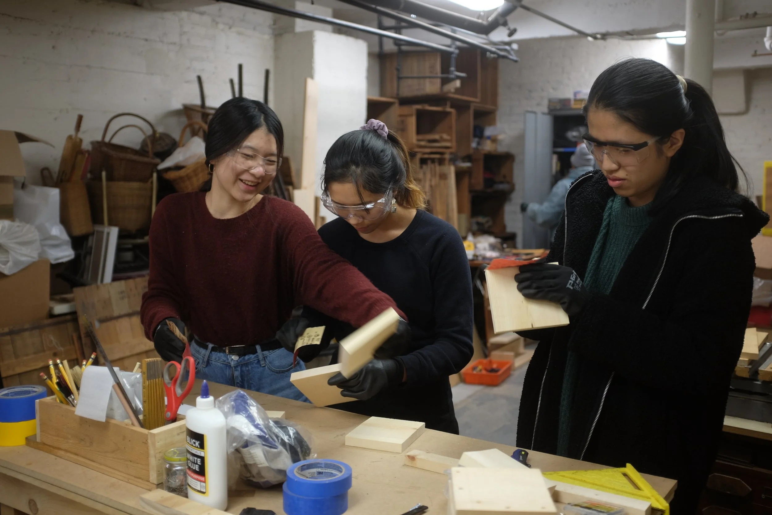 Three women working on woodworking projects in a workshop, wearing safety glasses and gloves, with various tools and wooden pieces on the table.
