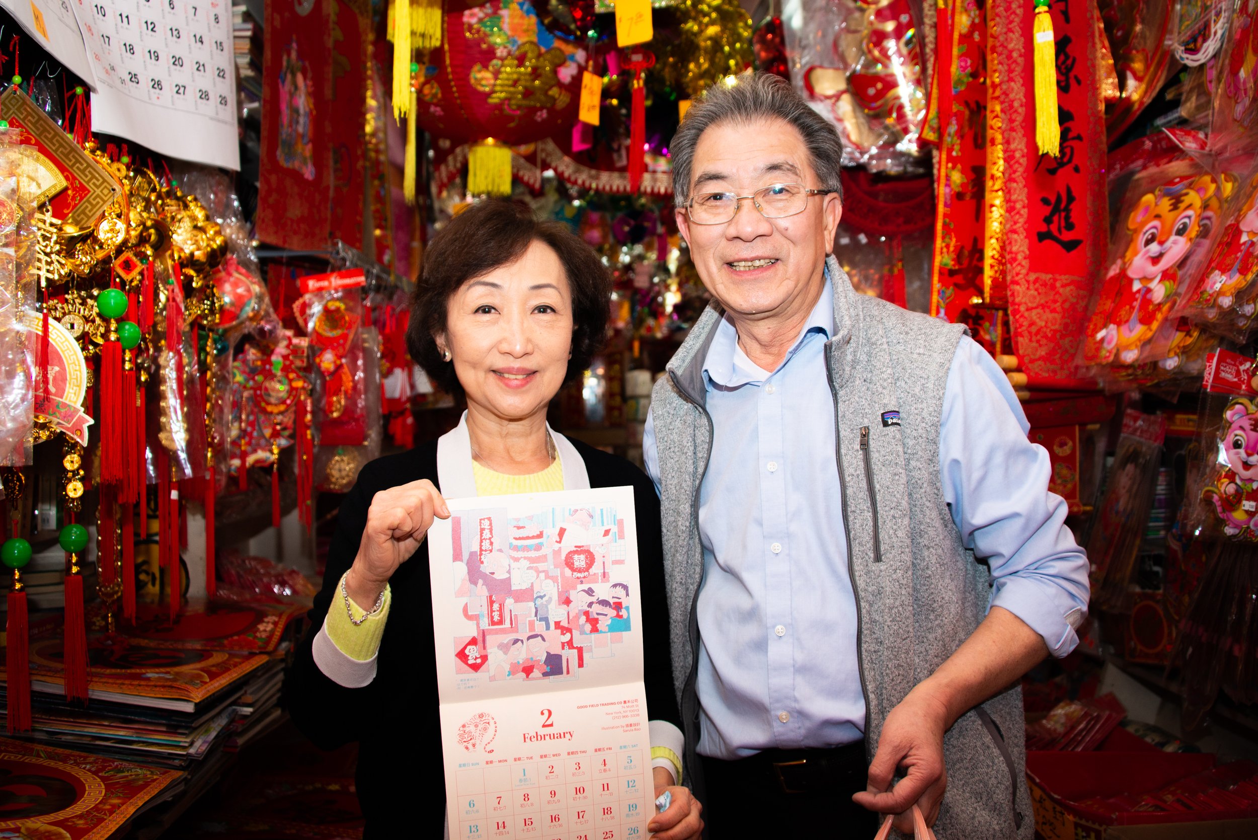 Two smiling elderly people, a woman holding a February 2023 calendar and a man standing next to her, in a shop decorated with traditional Chinese New Year decorations.