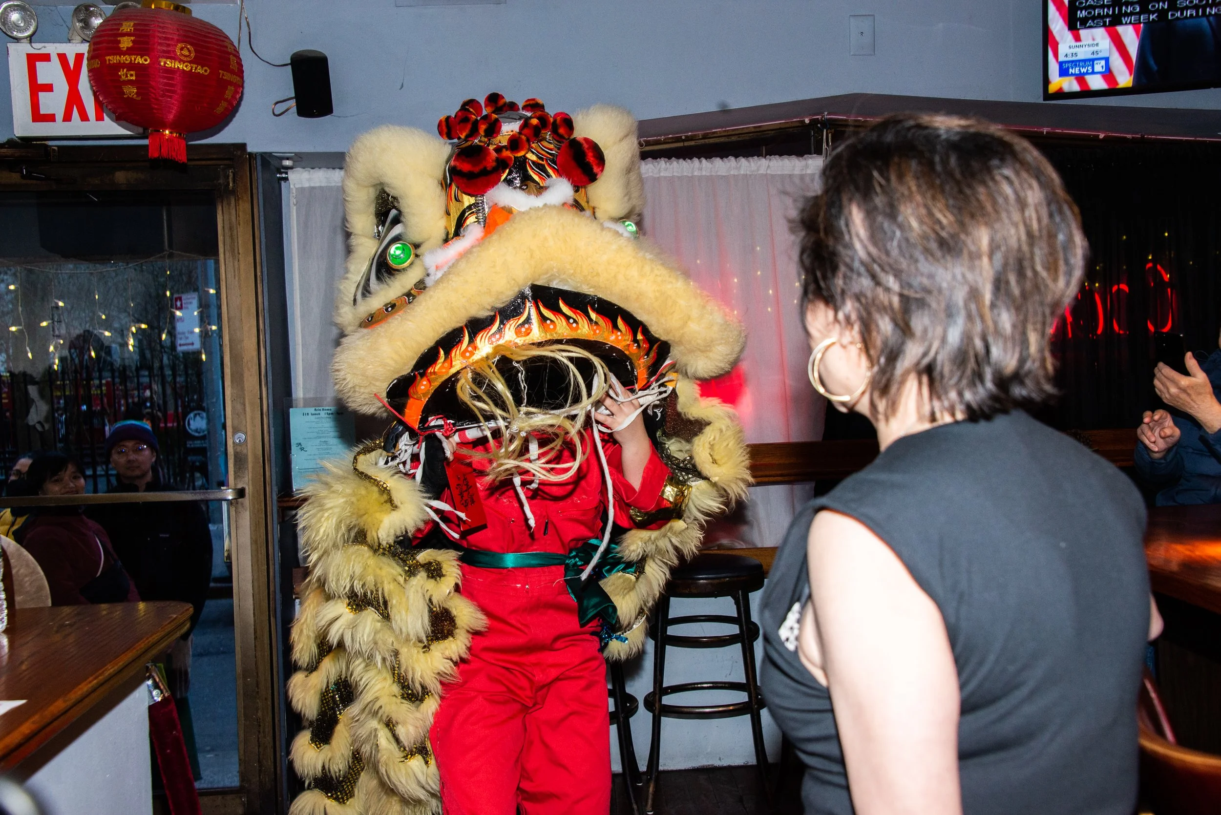 Person performing a traditional Chinese lion dance inside a restaurant, with the lion costume partially covering their face and wearing a red outfit; a woman with short brown hair and hoop earrings is watching the performance, and other patrons are visible in the background near the doorway.