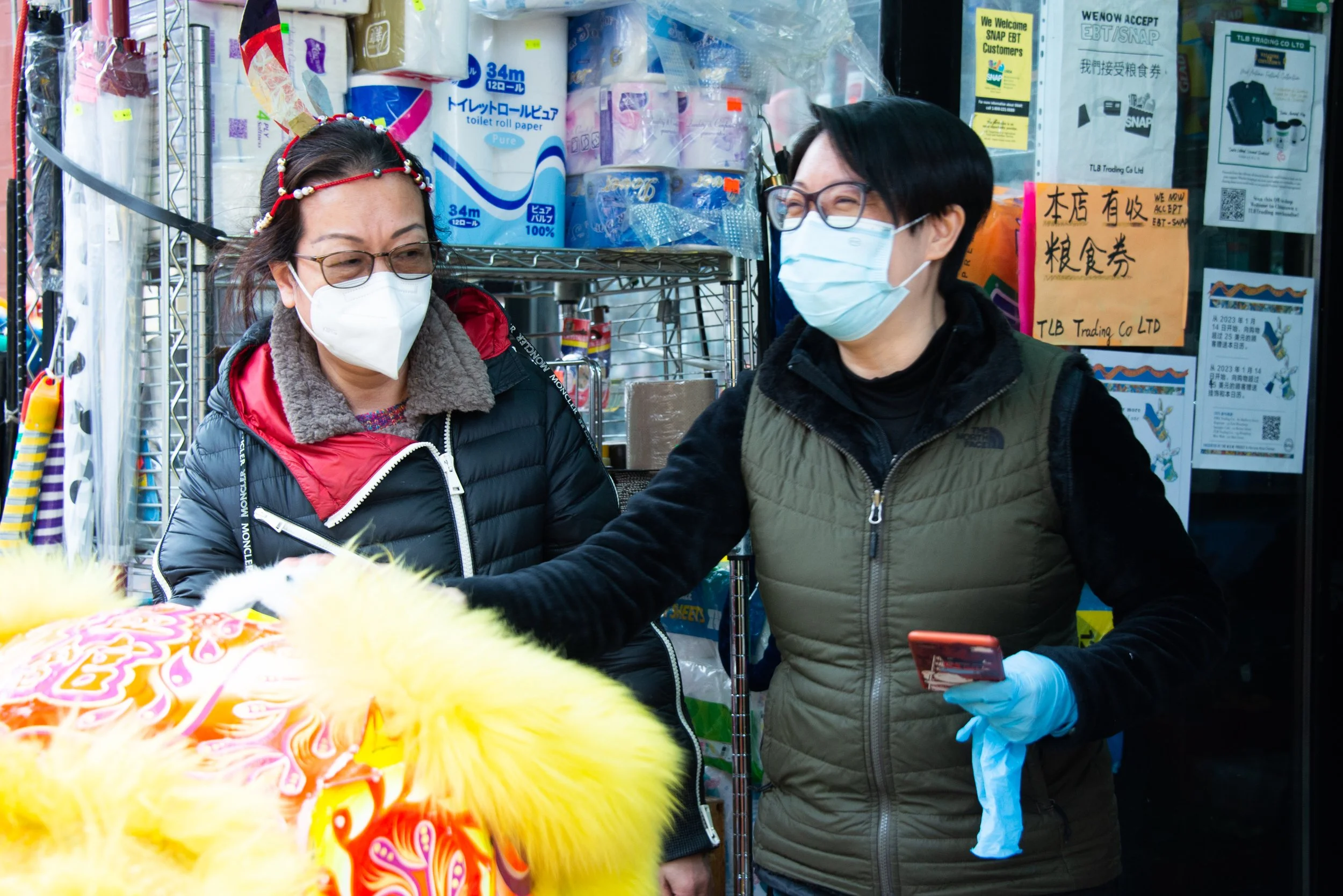 Two women wearing face masks in a store, one with glasses and a black puffy jacket, the other with glasses, black hair, and a green vest. They are in front of shelves with household supplies and signs in Chinese and English.