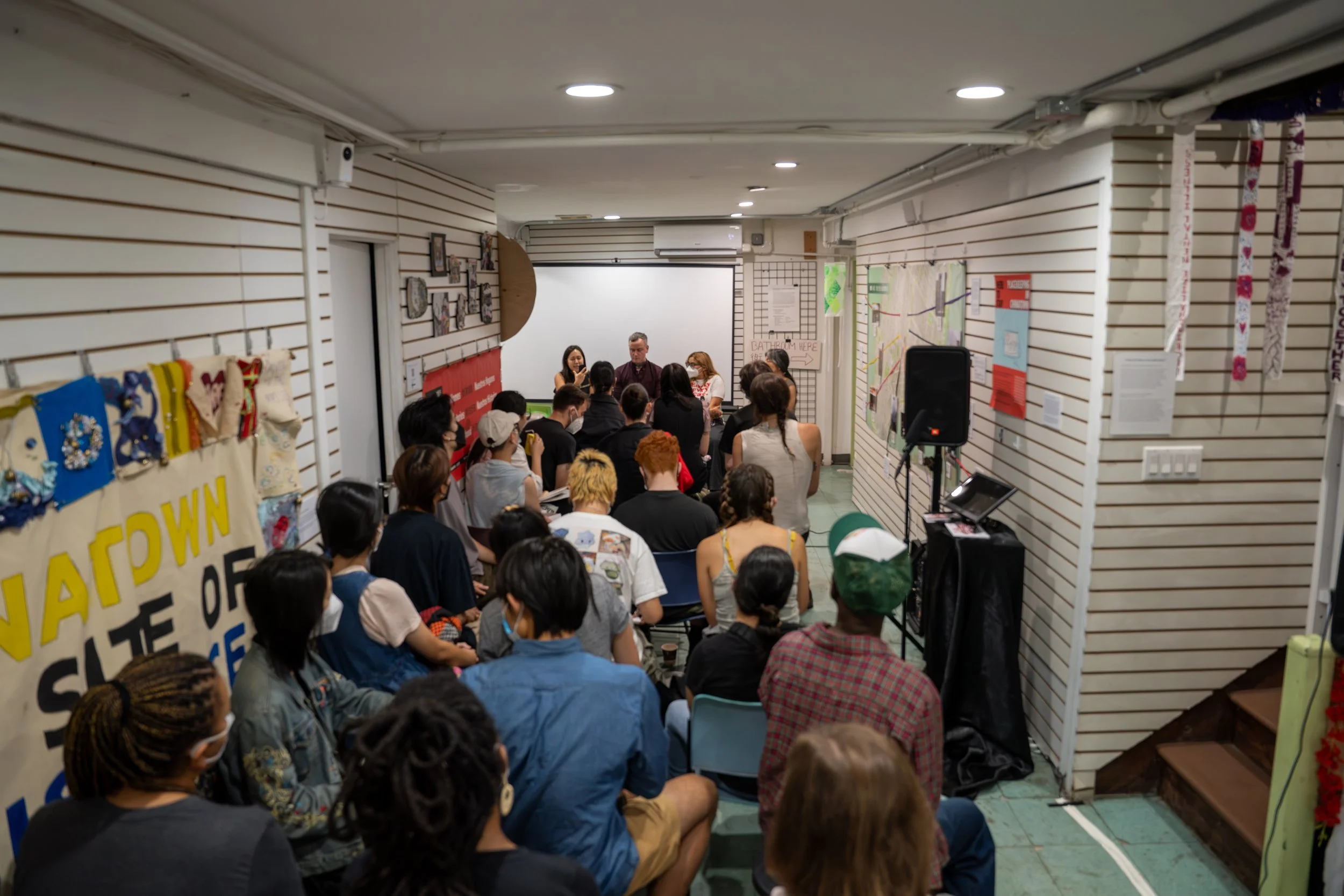 People attending a seminar or discussion in a room decorated with banners and artwork, with a panel of speakers at the front.