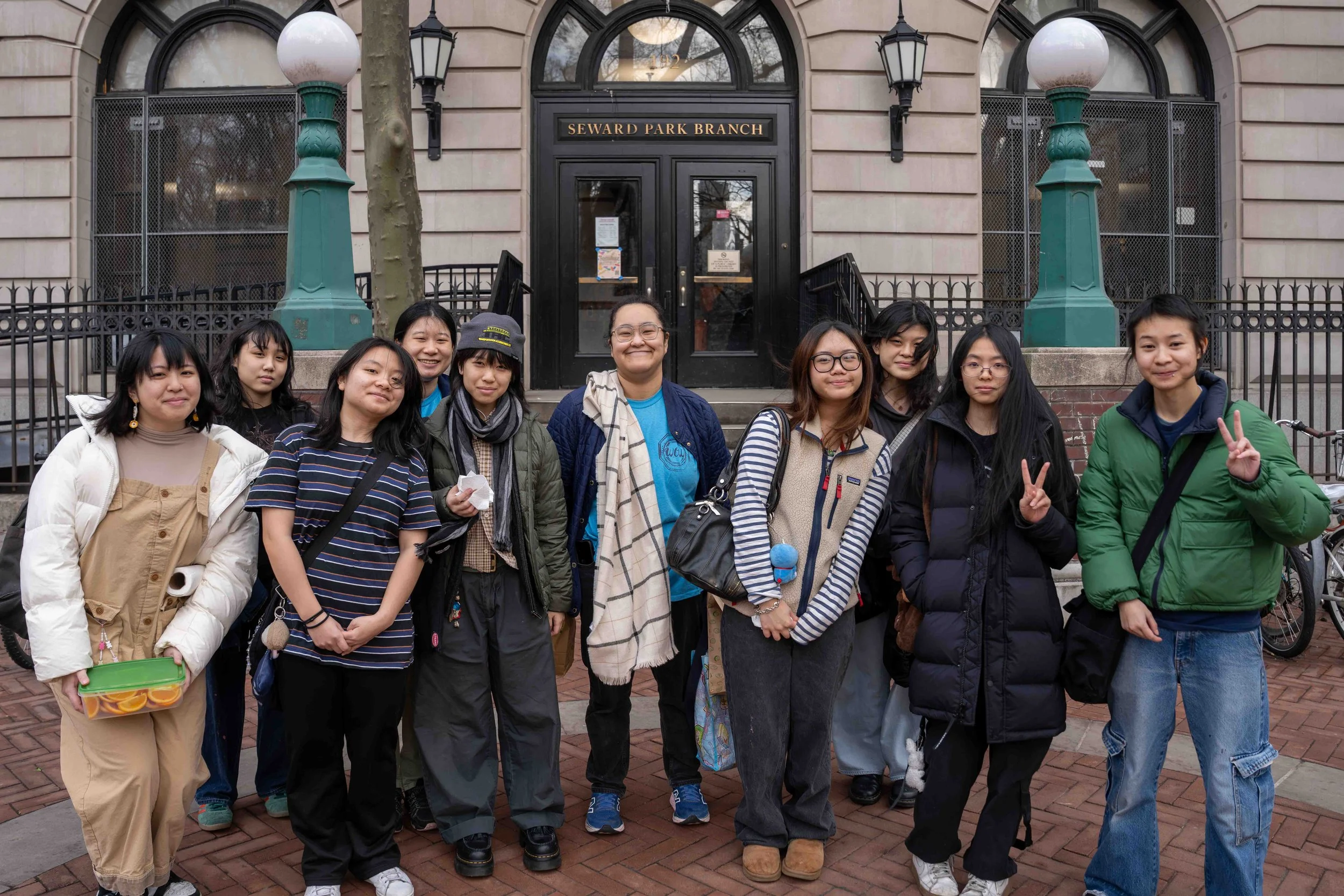 A group of nine young women standing outside Seward Park Branch with brick sidewalk, building entrance, trees, bicycles, and street lamps.