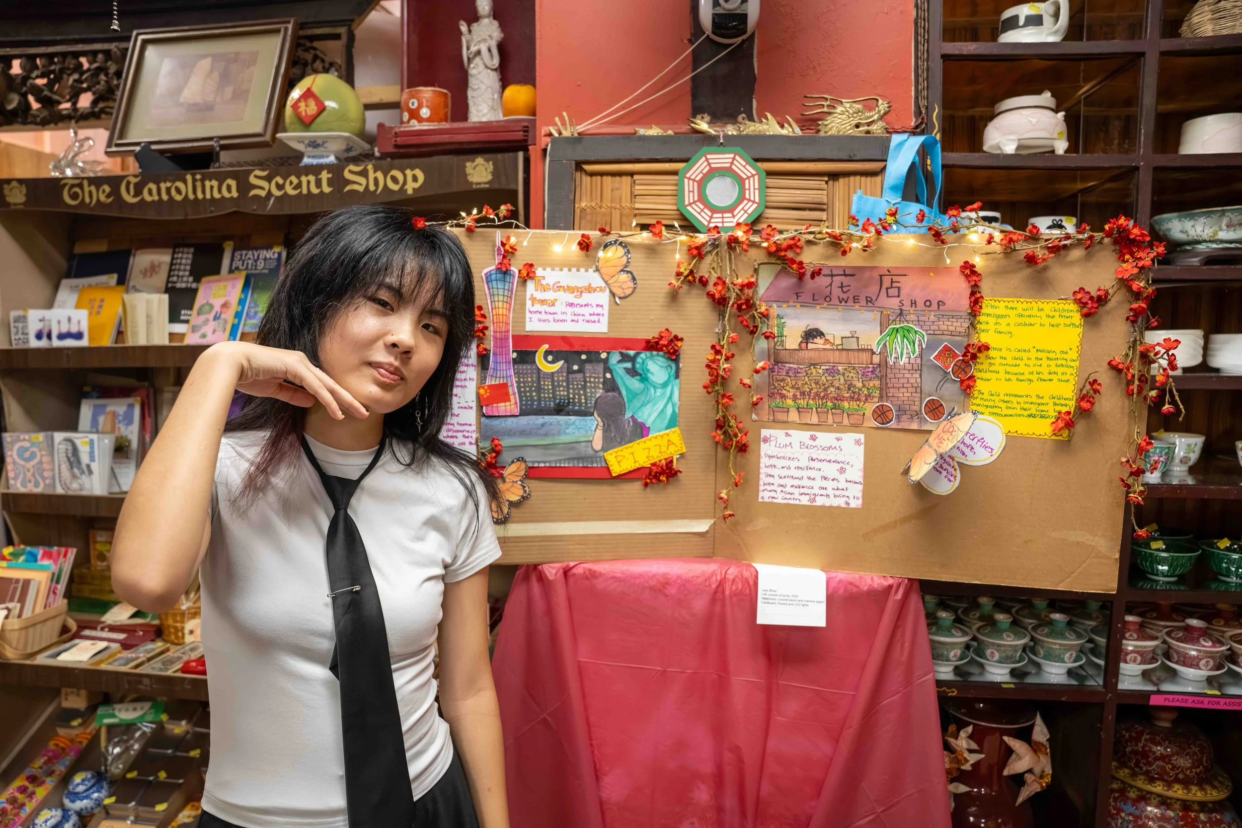 A young woman with black hair and a black tie posing in front of a display board adorned with red flowers, pictures, and handwritten notes inside a store named The Carolina Scent Shop, with shelves of ceramic bowls and kitchenware in the background.