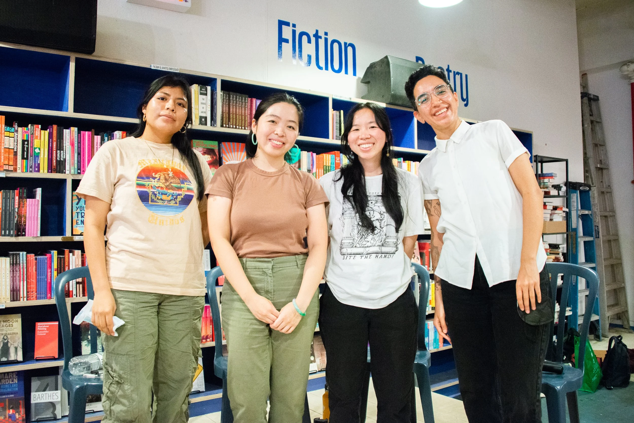 Four women standing together in a bookstore, smiling, with bookshelves labeled 'Fiction' and 'Poetry' in the background.