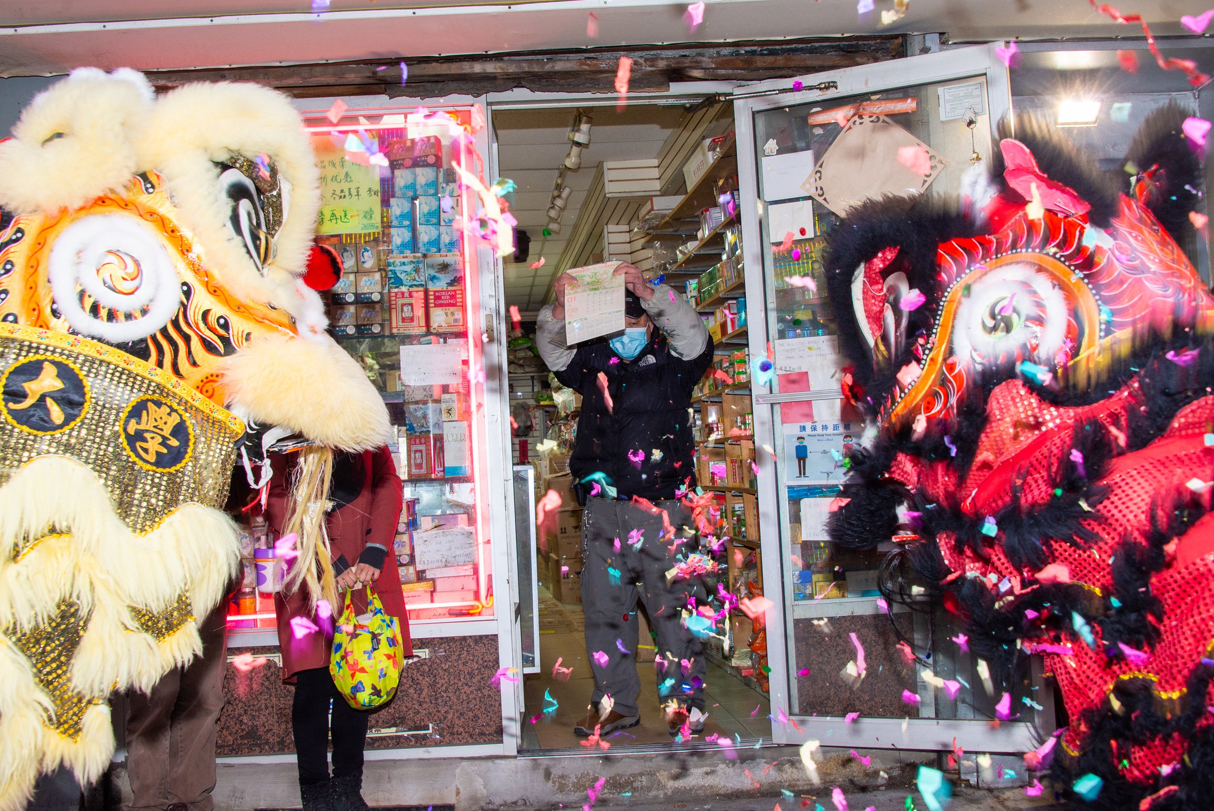 Two lion dance performers in traditional costumes performing inside a store, with a person wearing a face mask and holding a calendar in the middle, and colorful confetti falling through the air.
