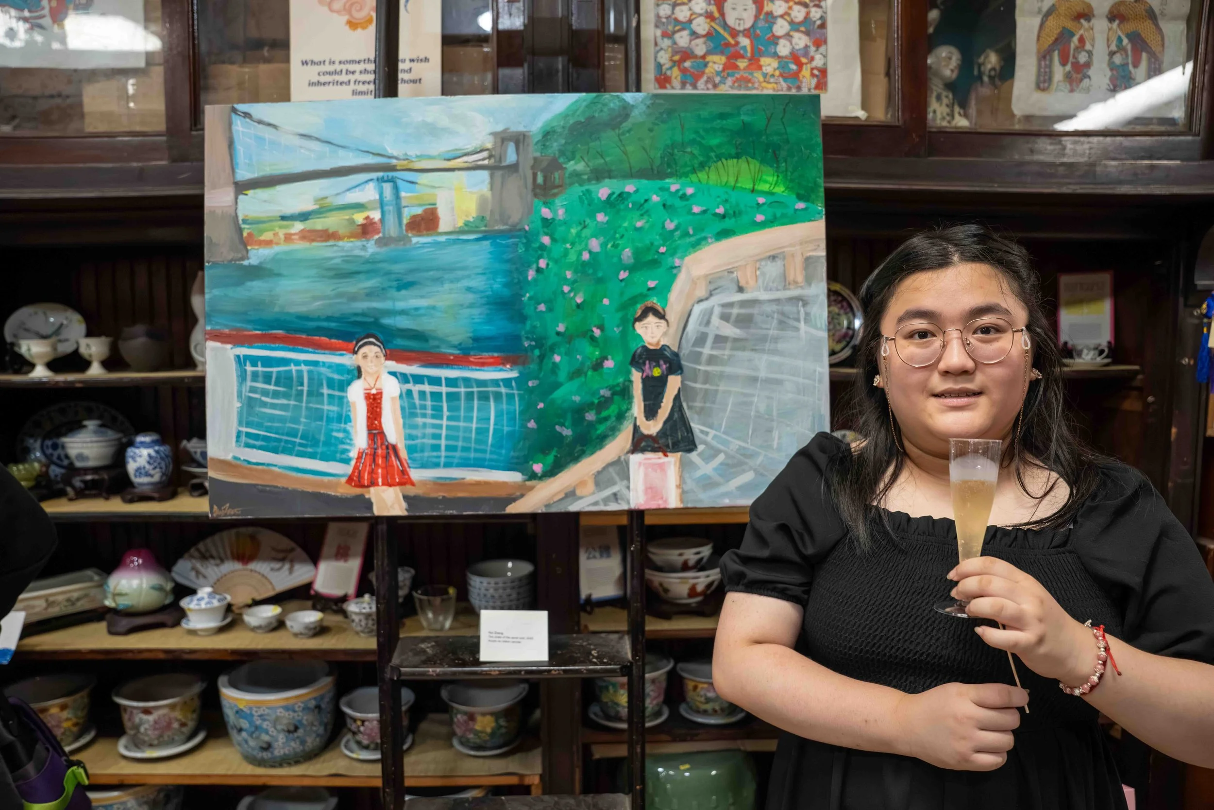A woman holding a champagne flute standing in front of a colorful painting of two girls, one at a sports field and the other on a garden path, with pottery displayed on shelves in the background.