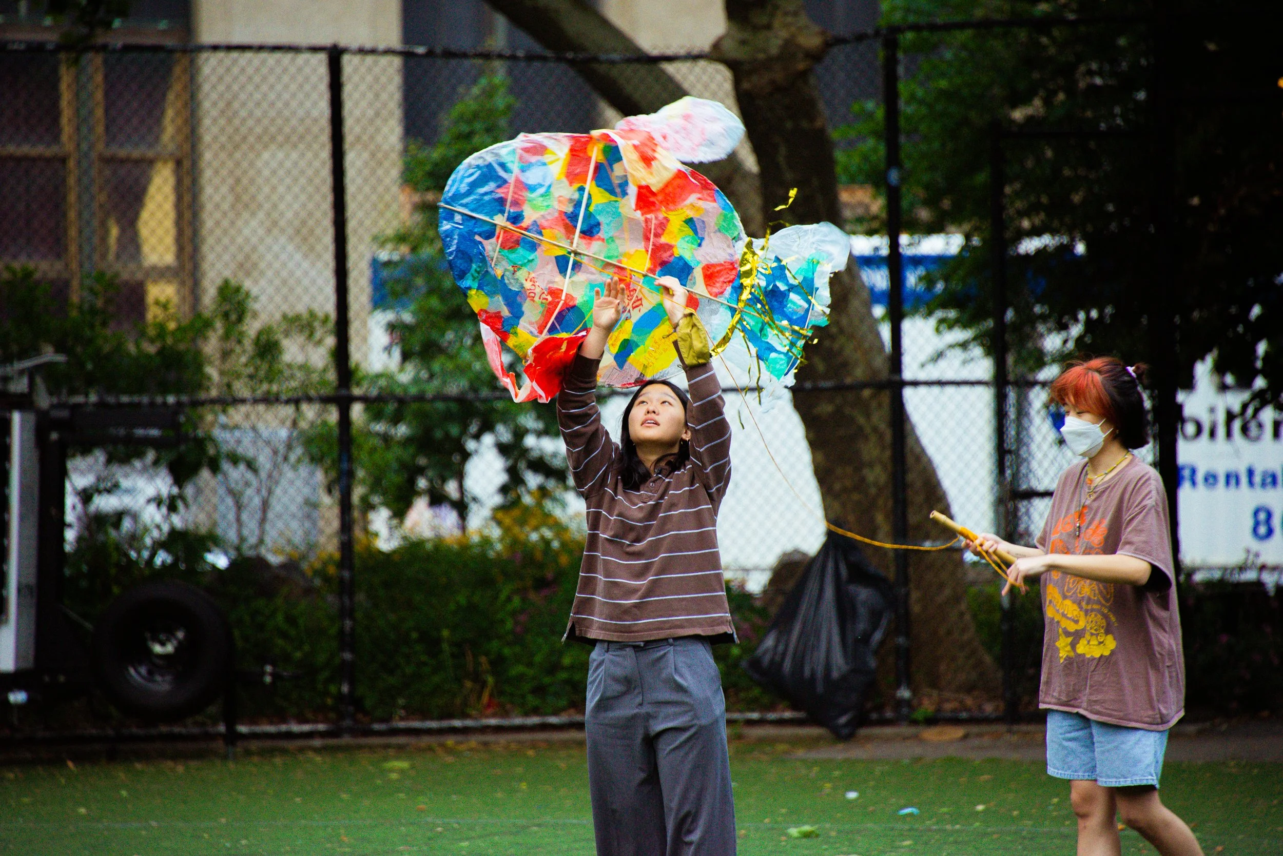 A girl flying a colorful kite outdoors, with another girl watching and holding a string, both wearing masks.