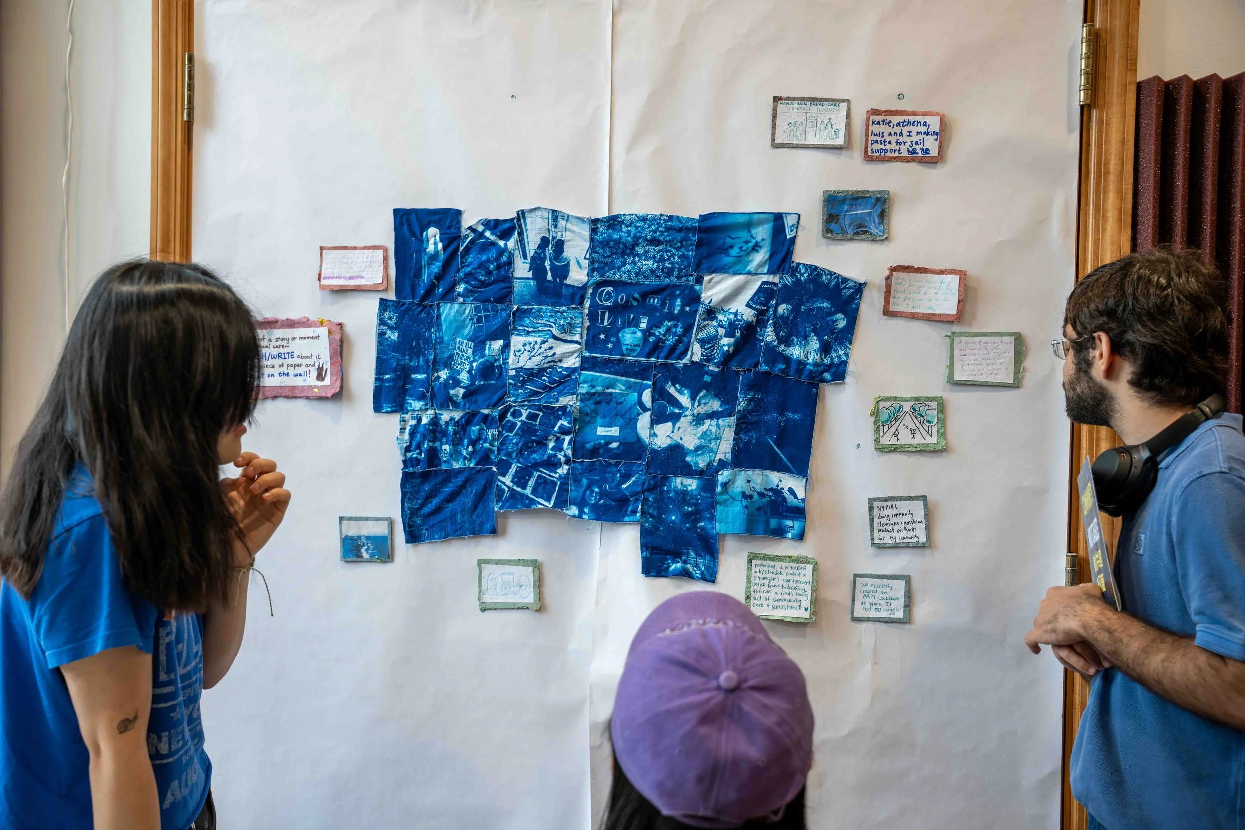 Three individuals observing an art display on a wall, which includes blue-toned images and handwritten notes surrounding the artwork.