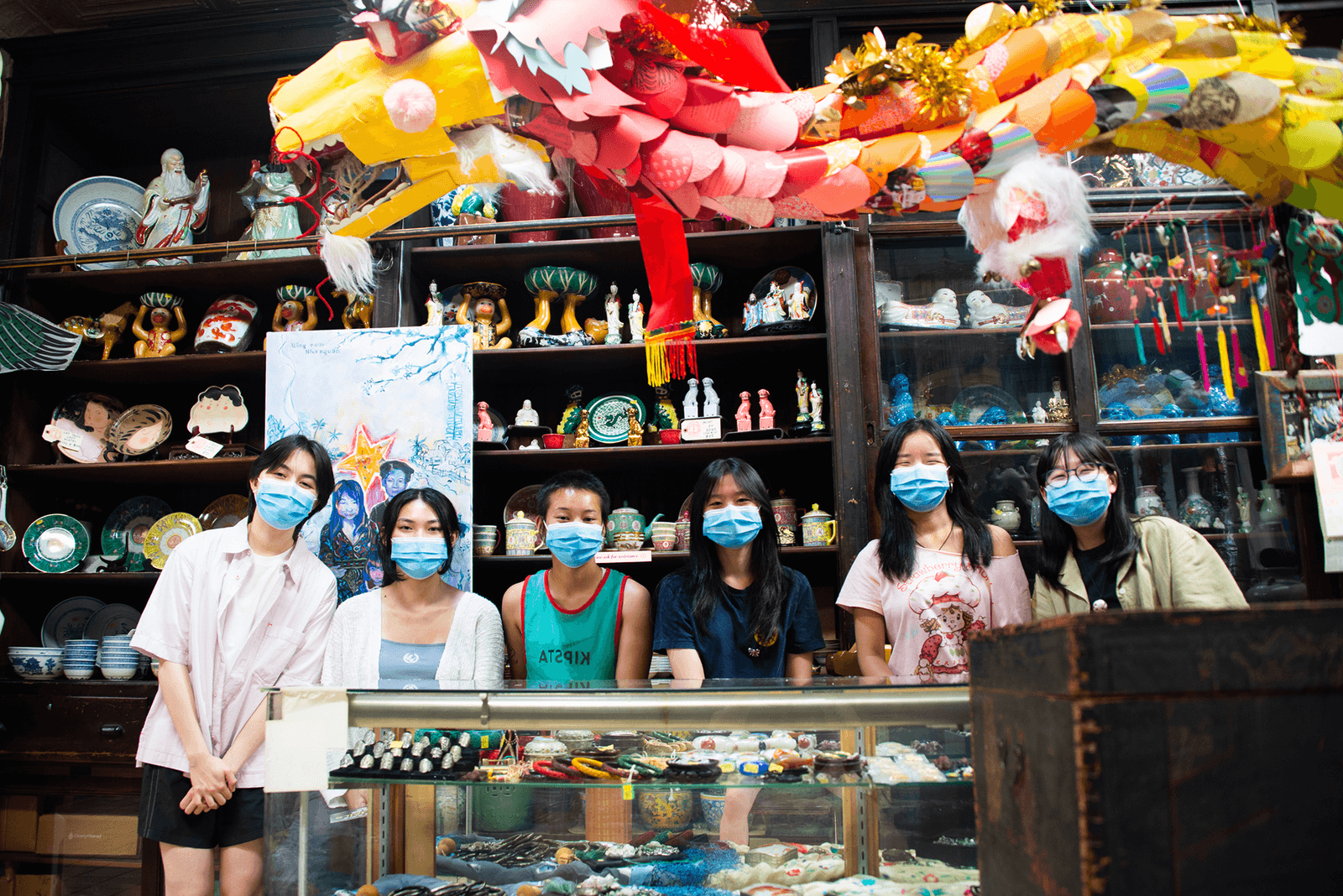 Five women standing behind a glass display case filled with jewelry, all wearing blue face masks, inside a shop with shelves of ceramics, decorative items, and colorful hanging decorations.
