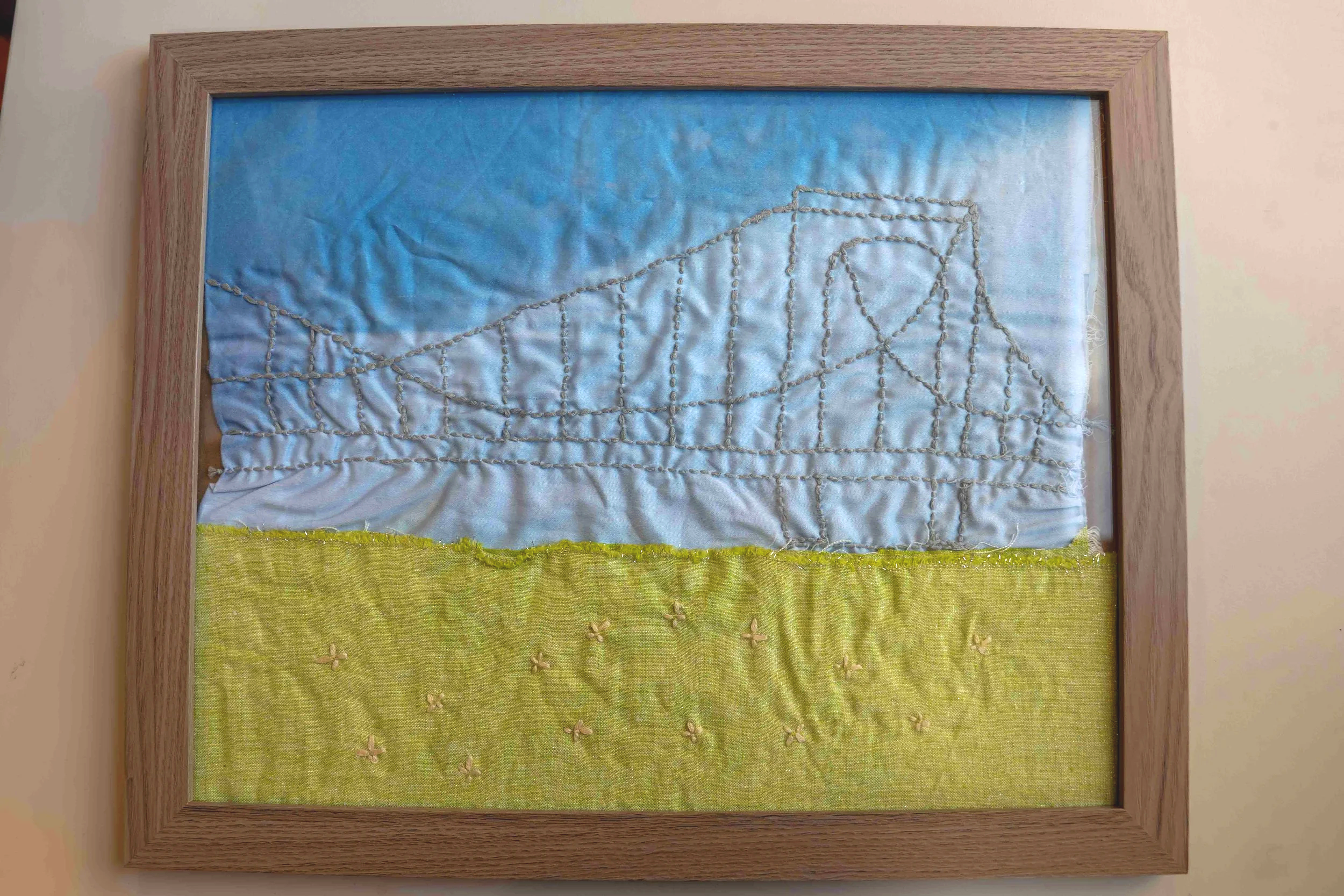Framed embroidered fabric artwork depicting a landscape scene with a blue sky and green field, embroidered with threads and small floral embellishments.