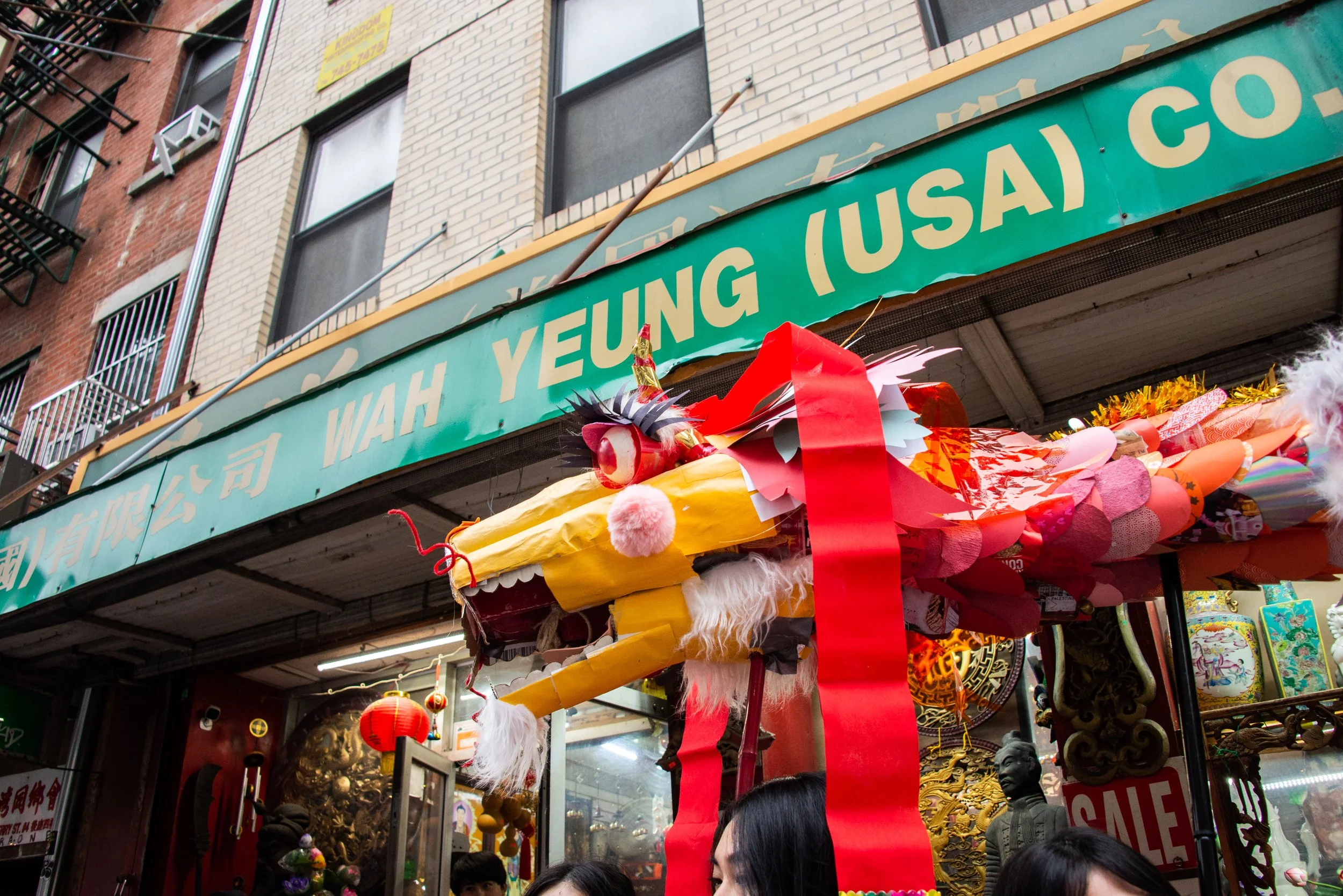 A Chinese dragon head decoration hanging outside Wah Yeung (USA) Co. store during a celebration or festival with red lanterns and people visible.