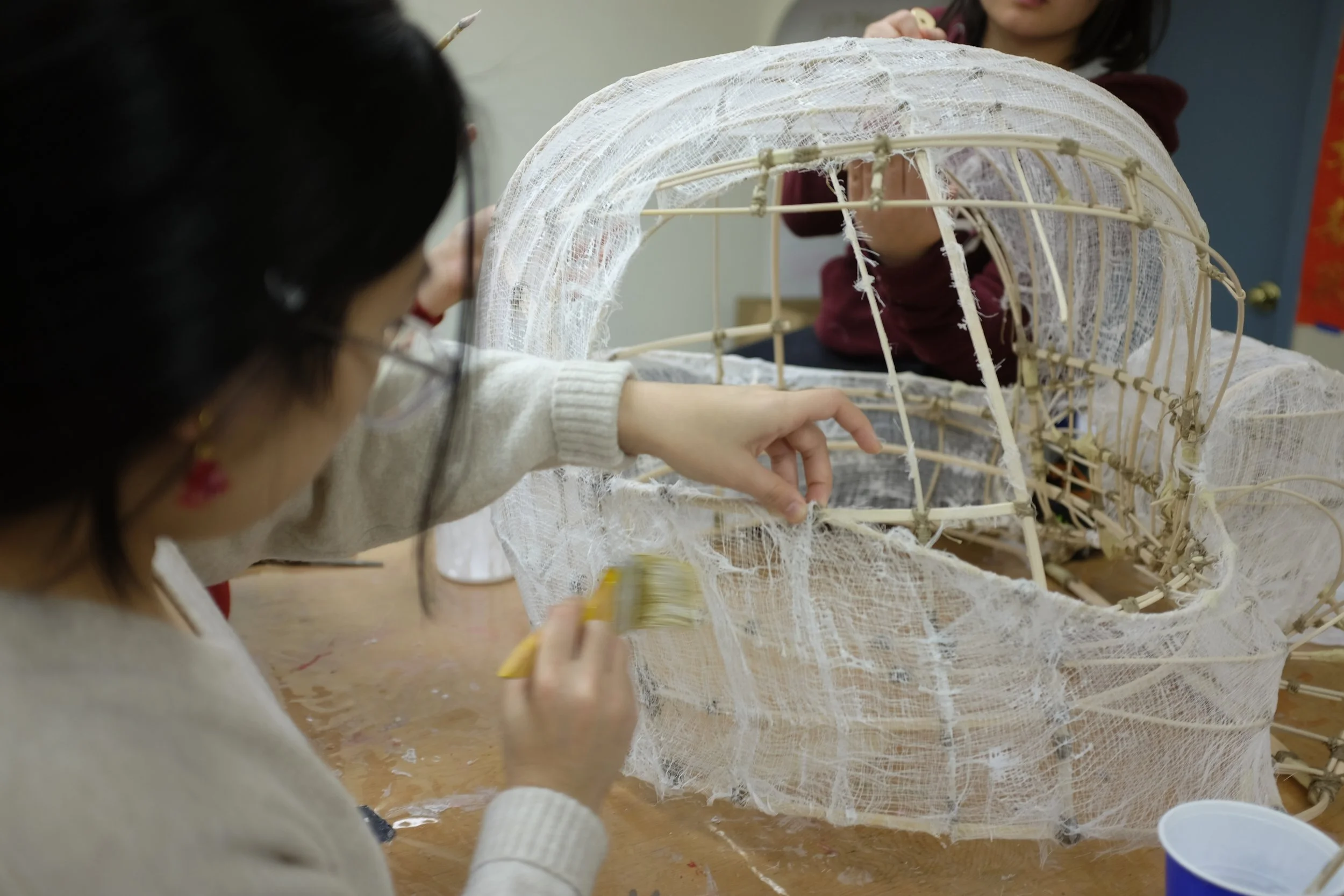 A woman working on a wire sculpture with a paintbrush, wrapping white fabric around it, in a workshop or classroom setting.