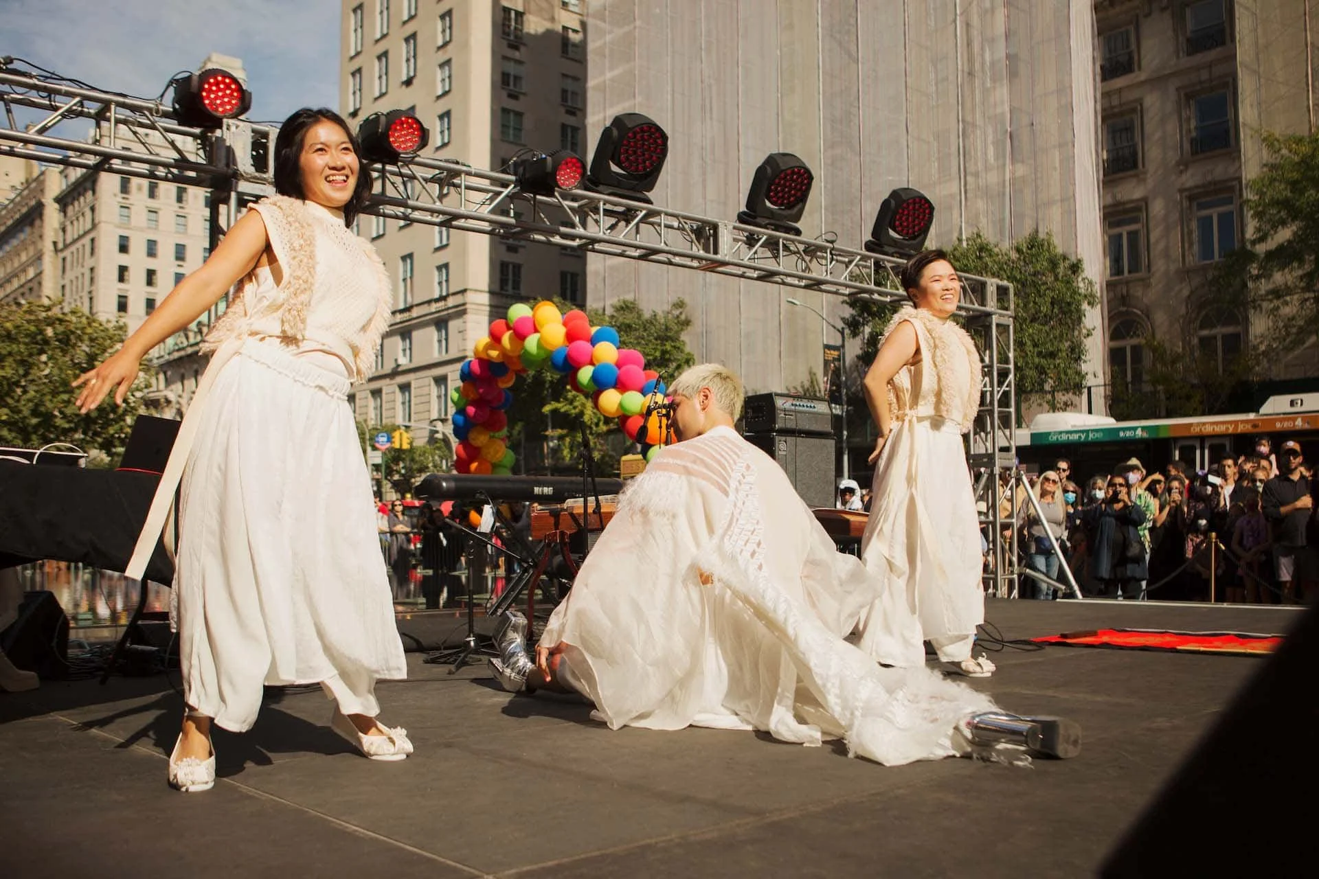 Three women in white dresses dancing on an outdoor stage in a city, with one woman kneeling and holding a shoe, and a colorful balloon arch in the background.