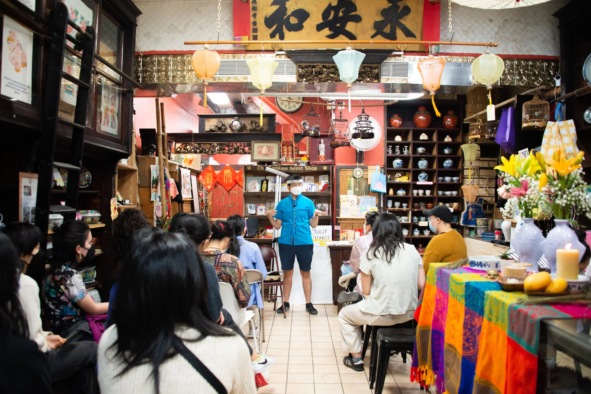 Group of people seated in a shop decorated with Asian ornaments, listening to a person in a blue shirt and white face mask speaking at the front.