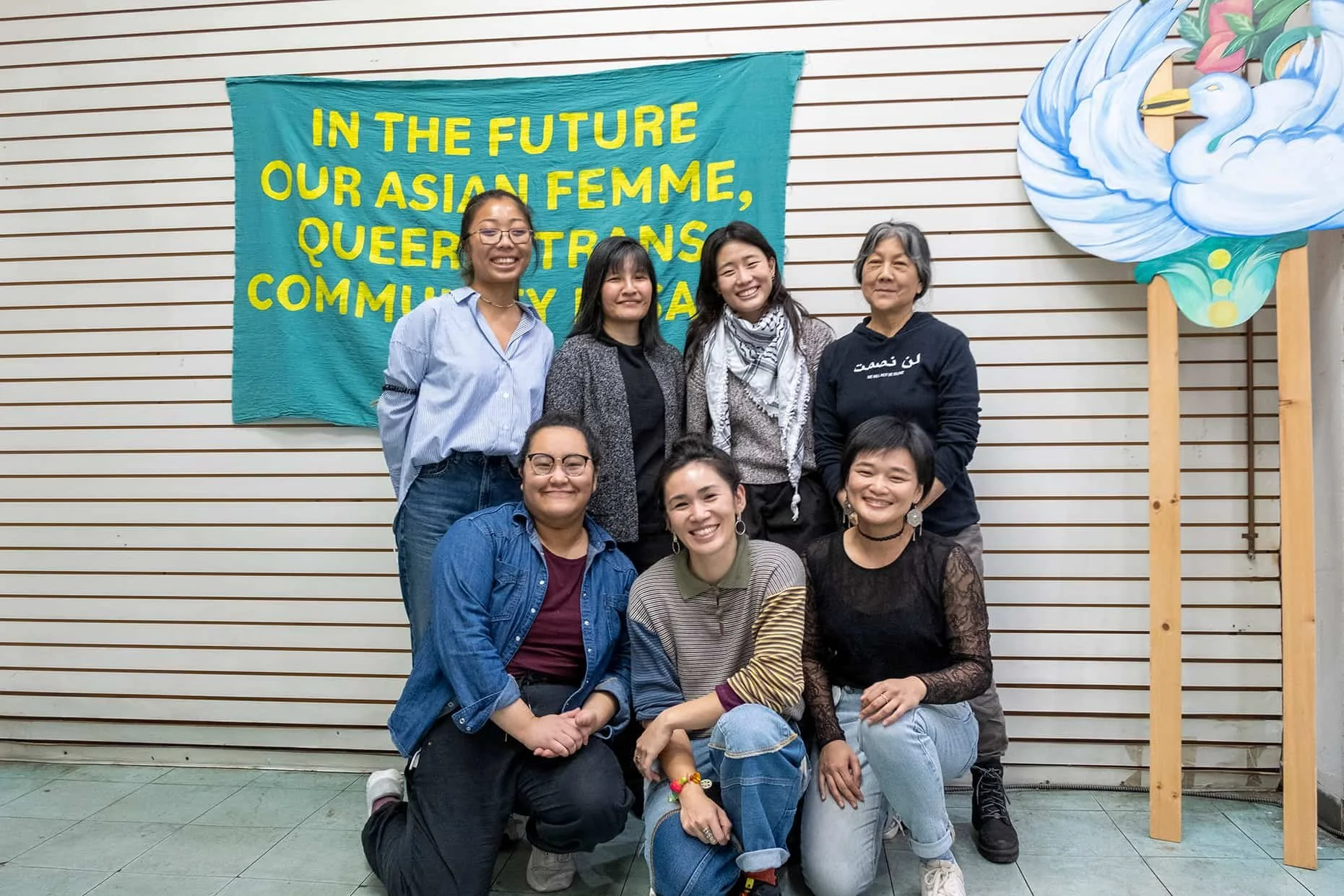 Group of seven women smiling and posing in front of a colorful banner that reads, 'In the future our Asian femme, queer trans community' and a swan artwork on the wall.