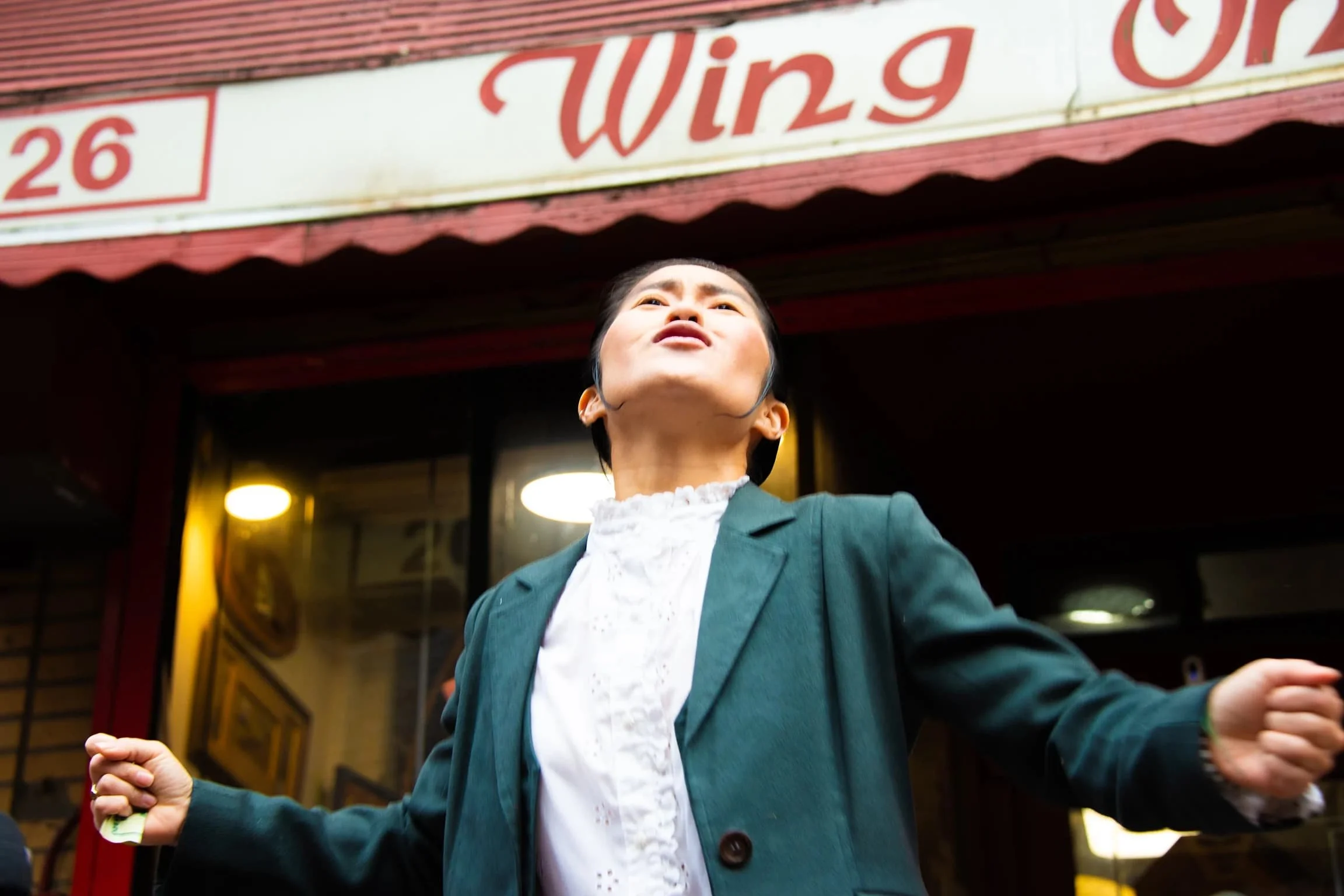 A woman with short black hair wearing a teal blazer and white blouse, standing outside a restaurant with a sign that says "Wing," gesturing with arms outstretched.