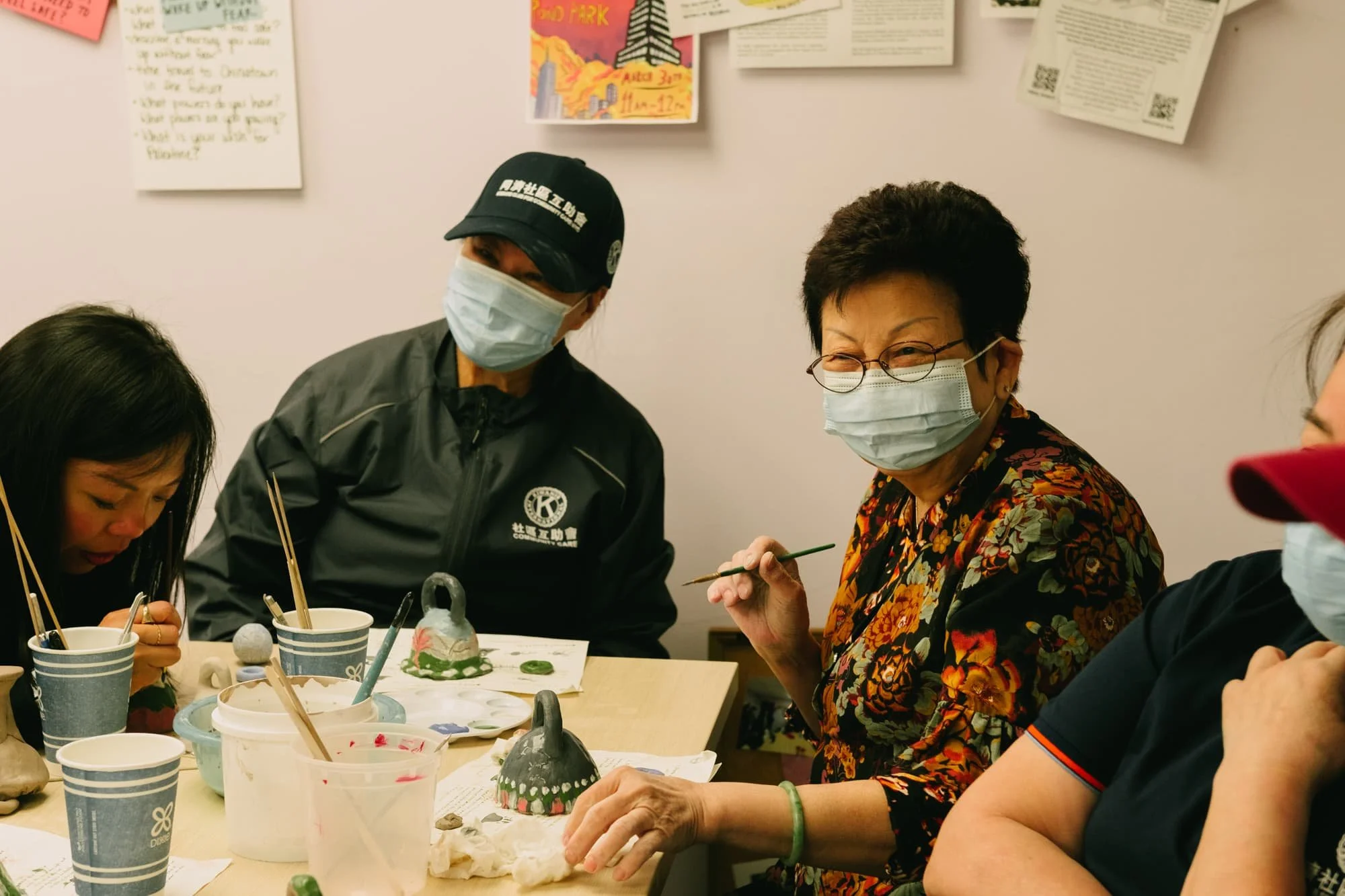 Group of four women sitting at a table, wearing masks, engaging in arts and crafts involving painted ceramic objects and brushes.