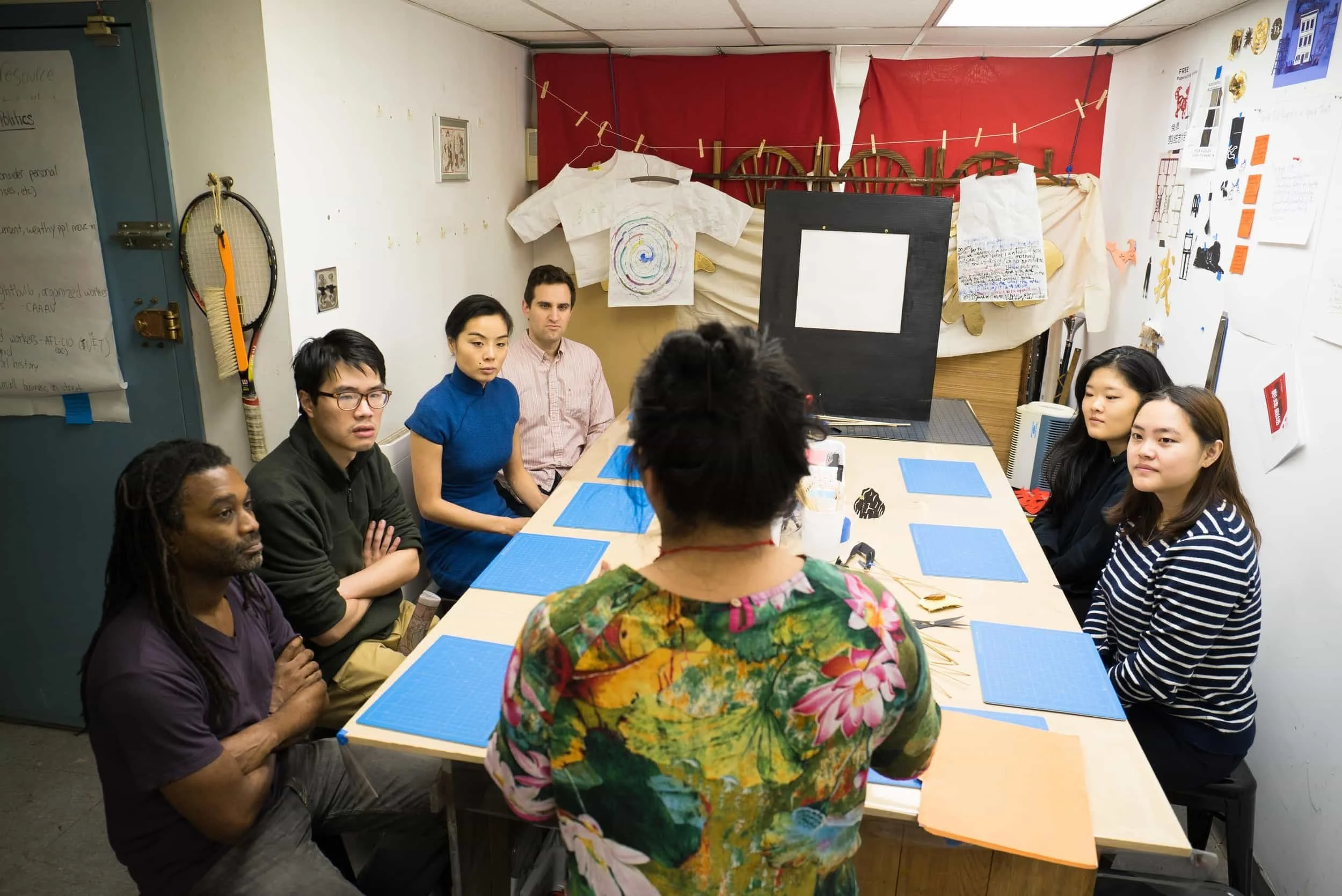 A group of seven people sitting around a rectangular table in a small room, facing a woman who is standing with her back to the camera. The room is decorated with art supplies, posters, and clothing hanging on a string. The table has blue mats, and the woman is wearing a colorful floral shirt.