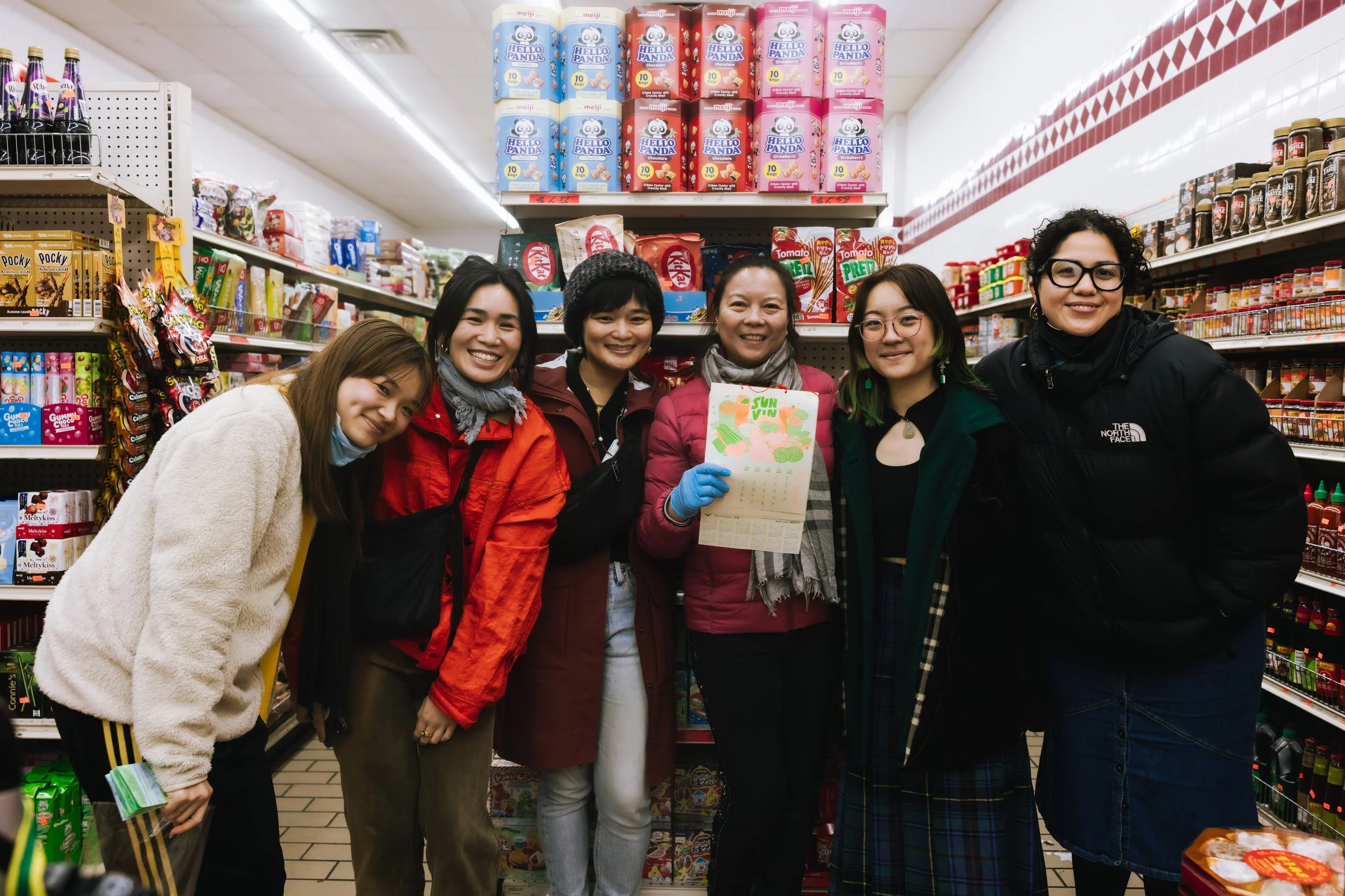 A group of six women standing together in a grocery store aisle, smiling, with snacks and products on shelves behind them.