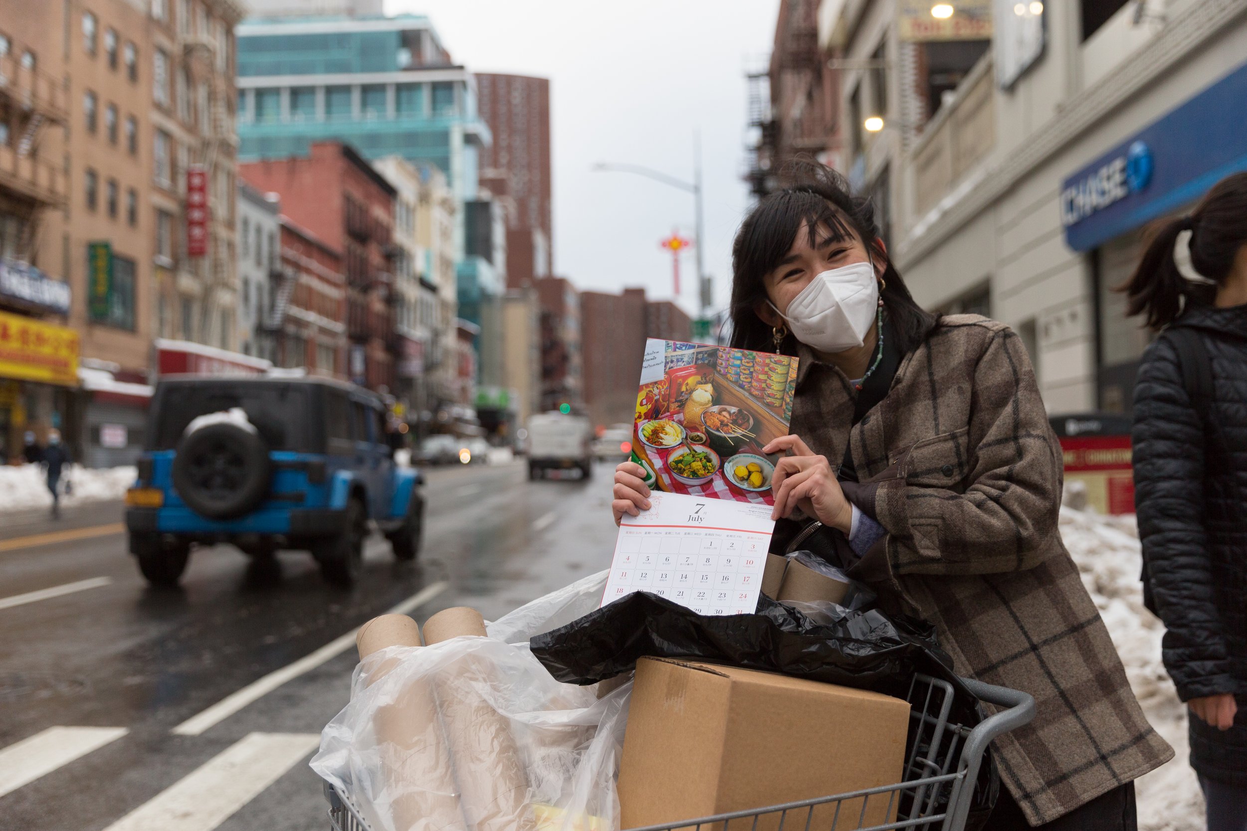 A woman holding a calendar showing July with a picture of food, standing with a shopping cart filled with boxes and bags, on a city street with buildings, cars, and snow.