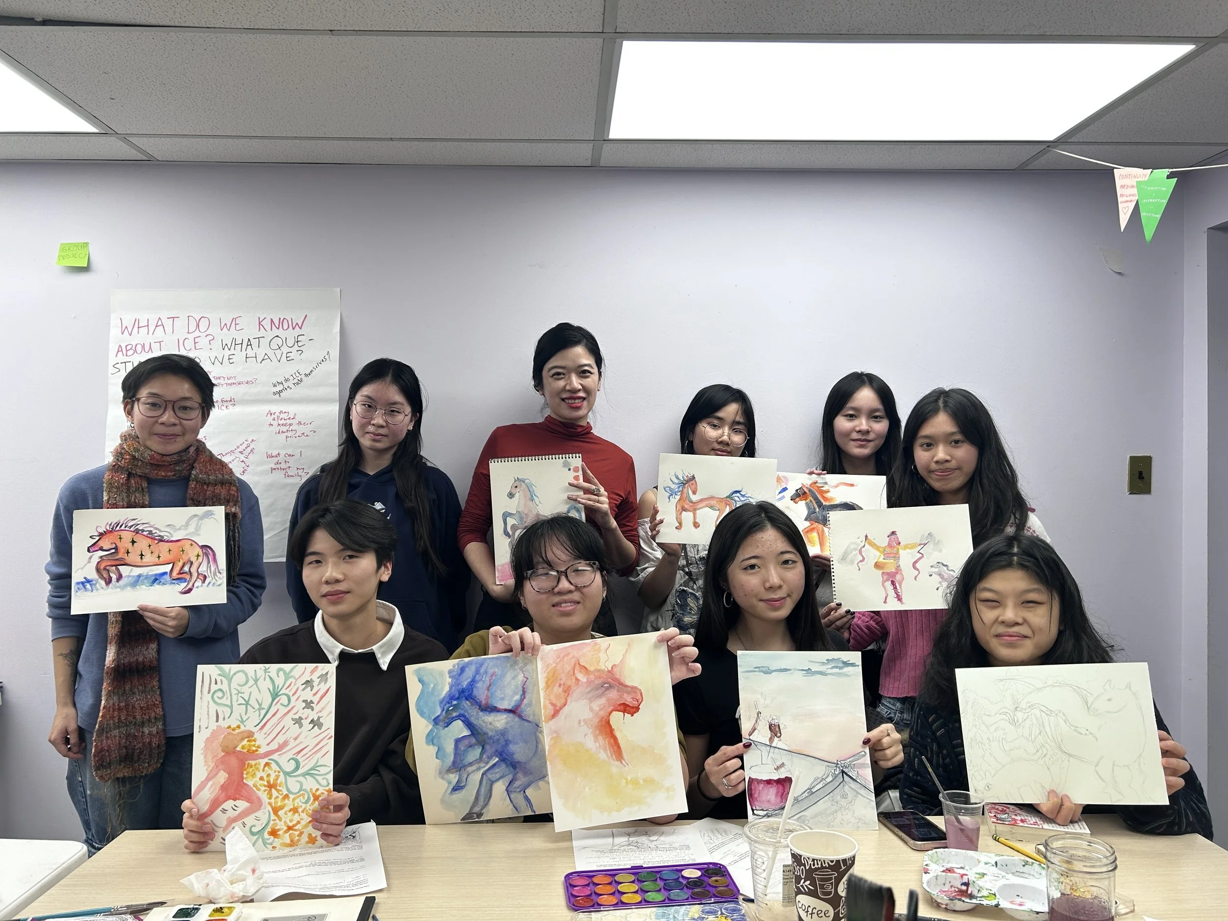 Group of ten women in an indoor setting holding up colorful animal watercolor paintings. Some are sitting at a table with art supplies and cups, others stand behind. There is a whiteboard with writing on the wall behind them.