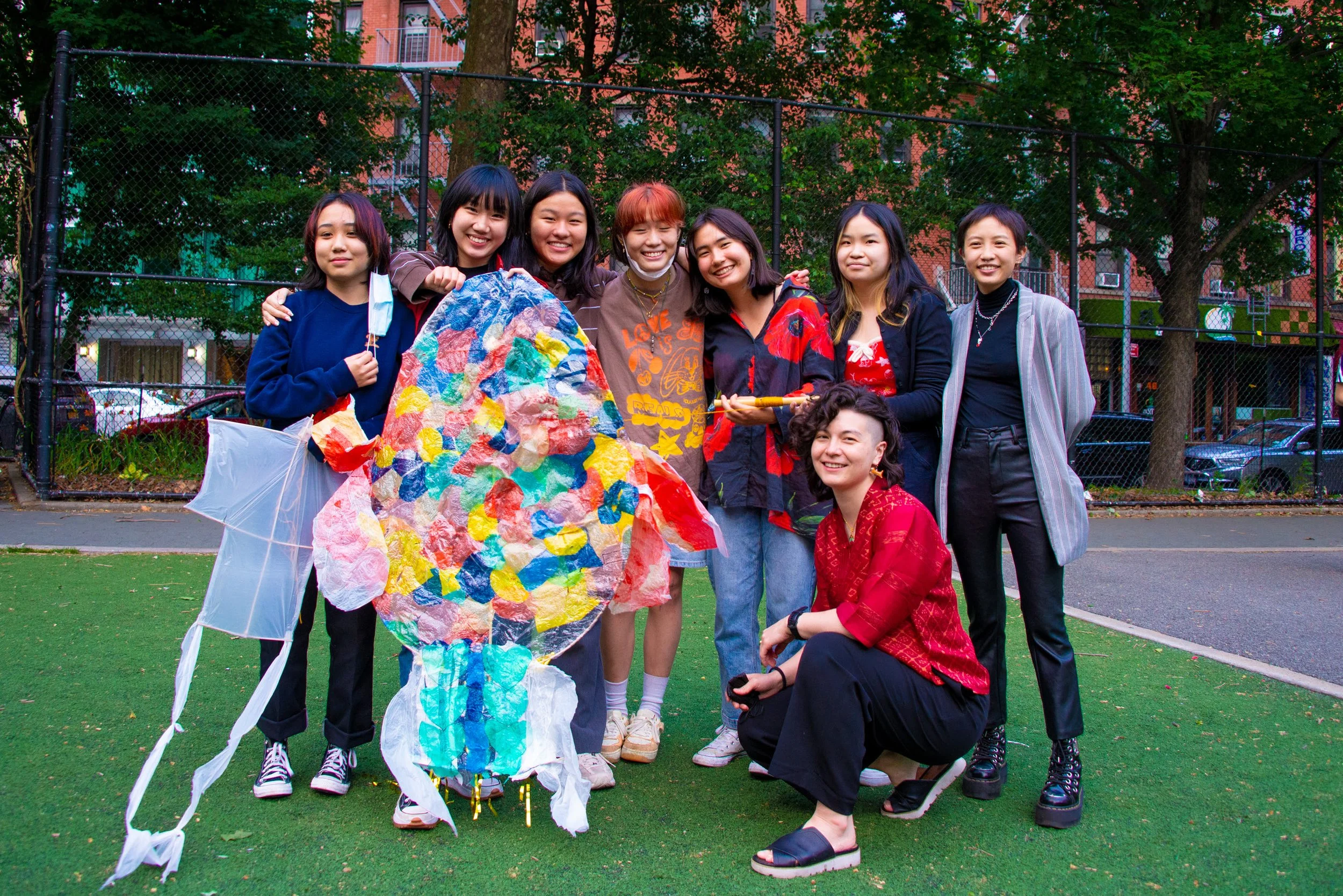 Group of nine young women standing together outdoors on a grassy area, smiling, with a colorful kite and paper lanterns, in front of a chain-link fence and trees, in an urban setting.