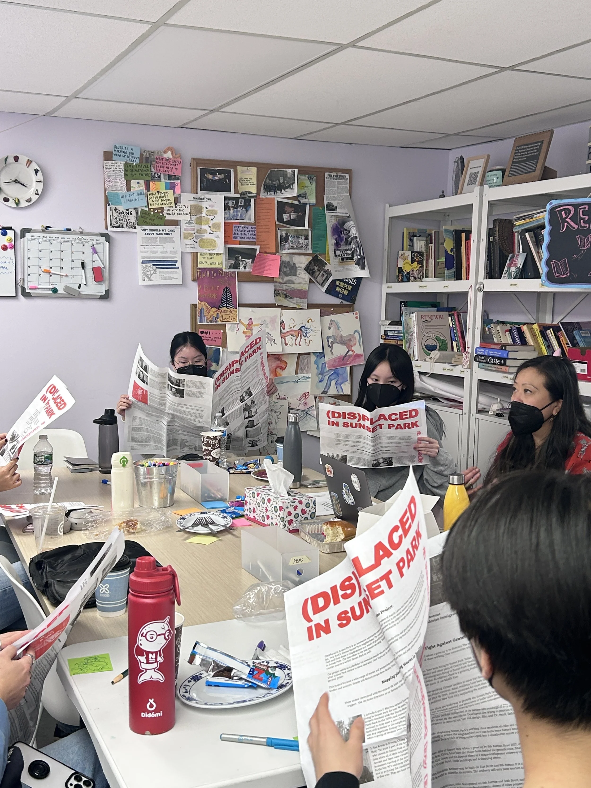 Group of people sitting around a table in a room, reading newspapers with the headline "DISPLACED IN SUNSET PARK." The room has bulletin boards with photos and notes, and bookshelves filled with books.