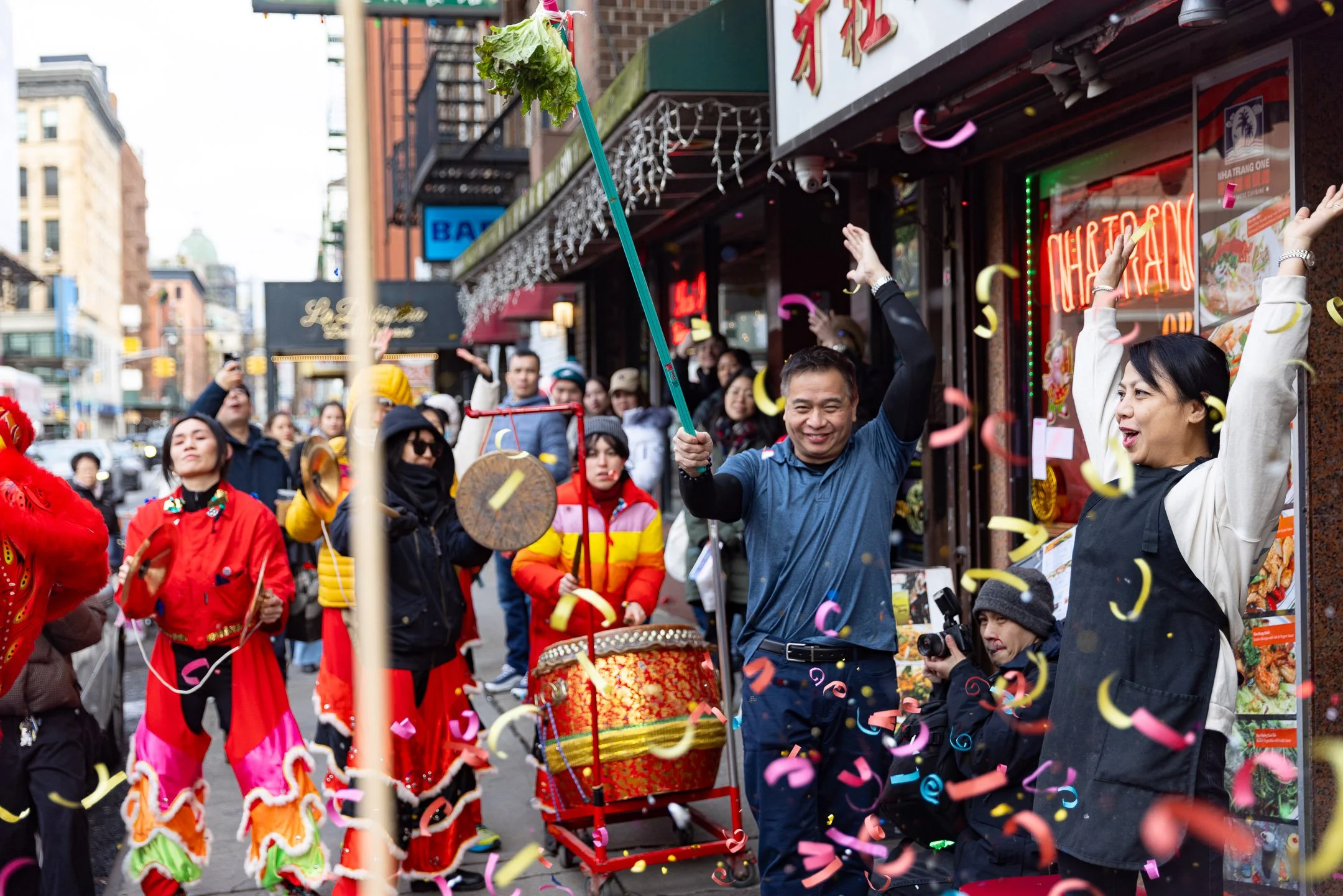 People celebrating on the street with confetti, some in traditional costumes, some with musical instruments, in front of a restaurant with neon signs and decorative lights.