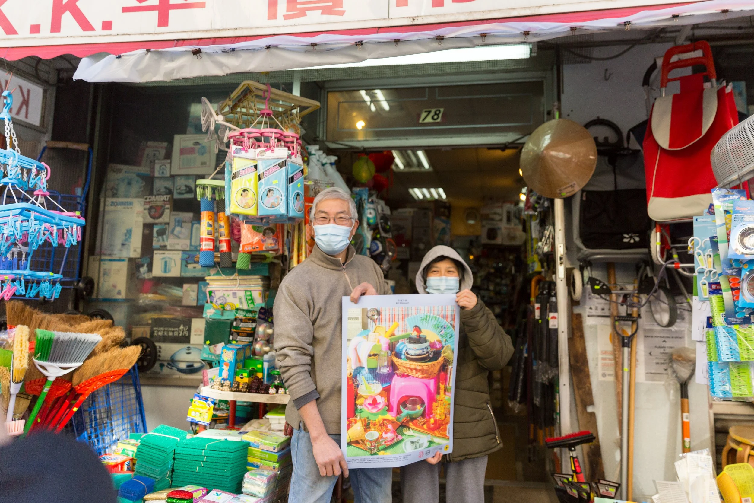 A man and woman wearing masks stand outside a street stall filled with various goods, holding a colorful poster.