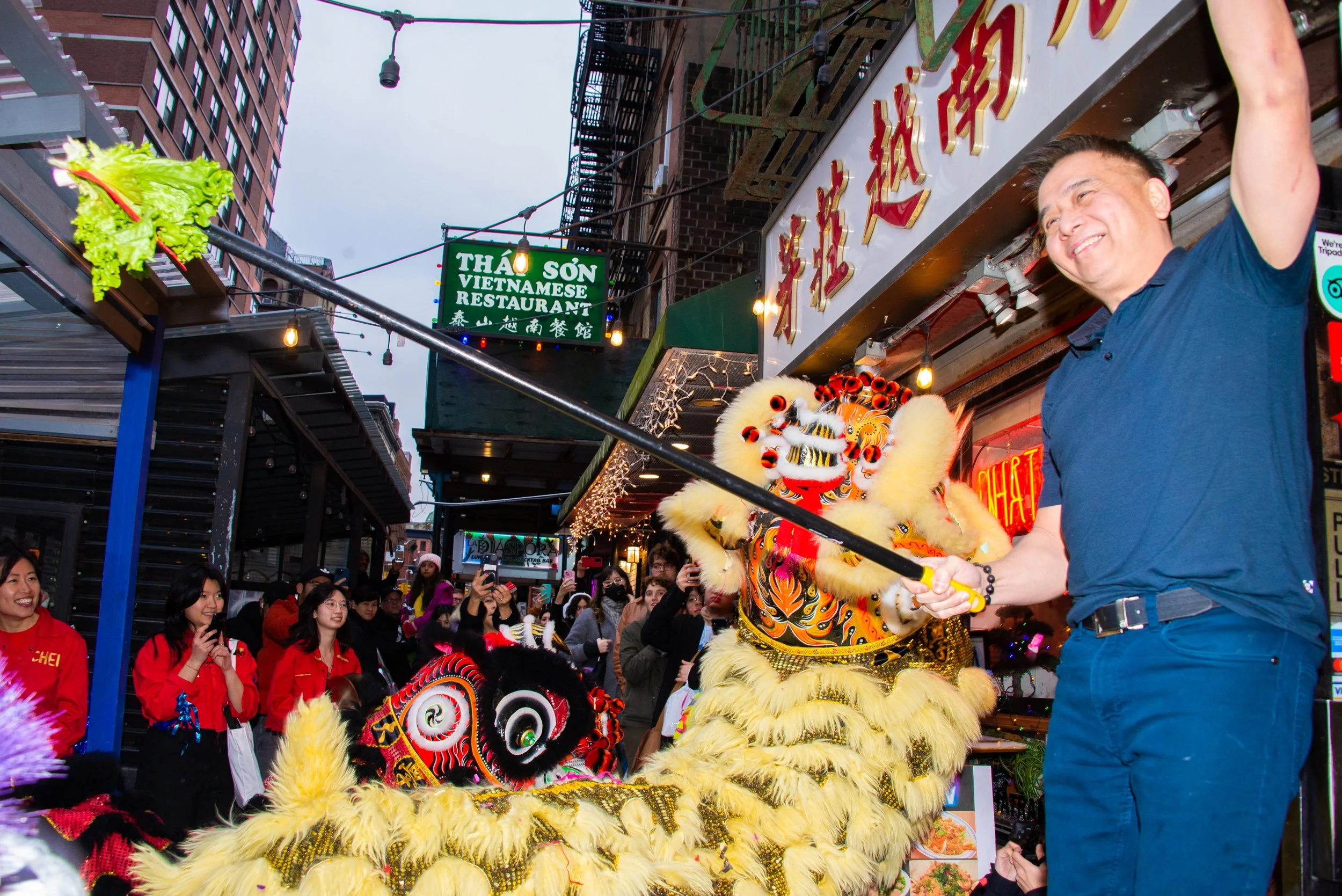 A man with a blue shirt holding a stick plays with a lion costume during a street parade, surrounded by onlookers in Chinatown.