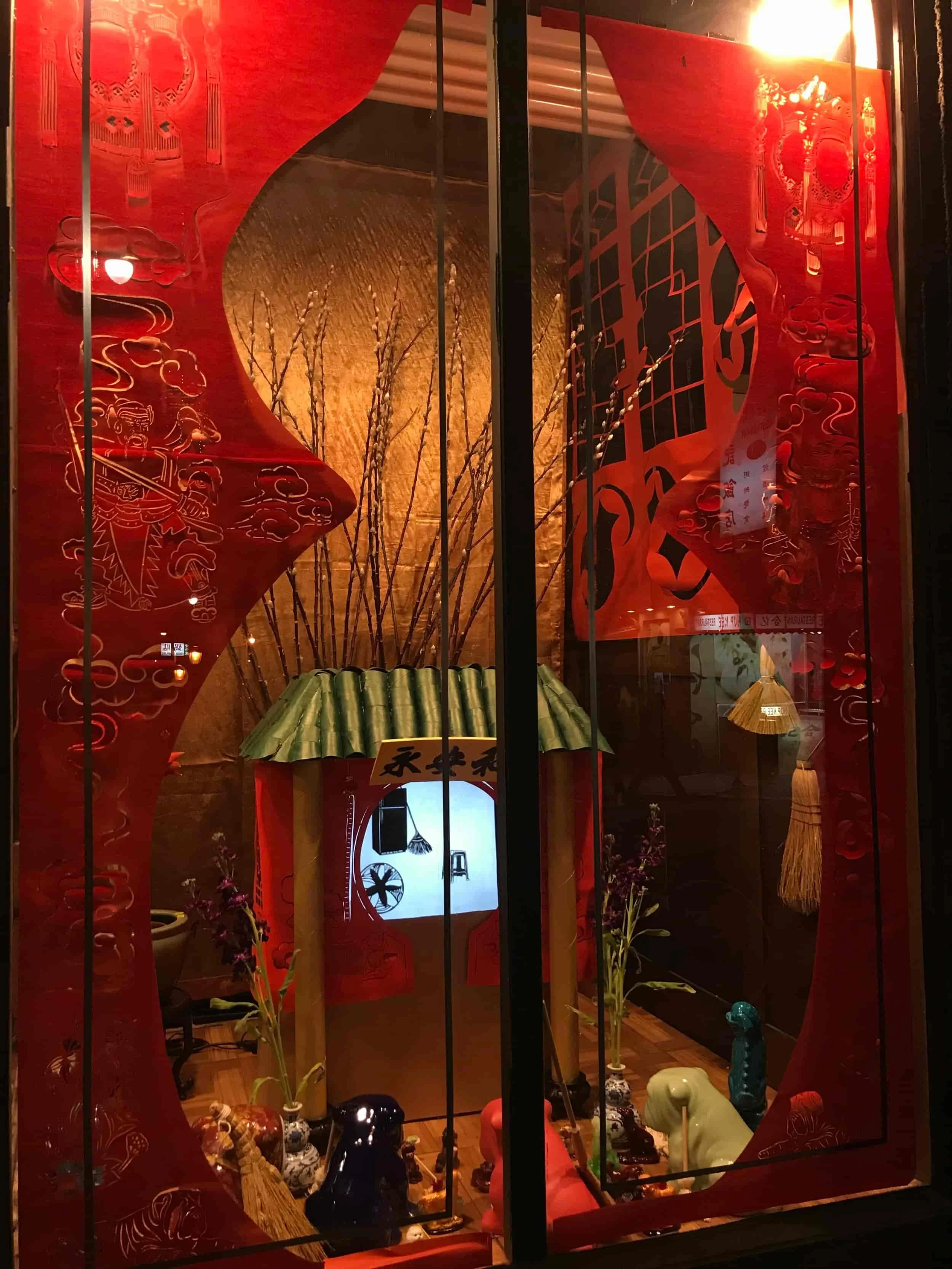 An Asian-inspired decorative altar with a red backdrop, featuring a small shrine with a green roof, vases with flowers, and various animal figurines.
