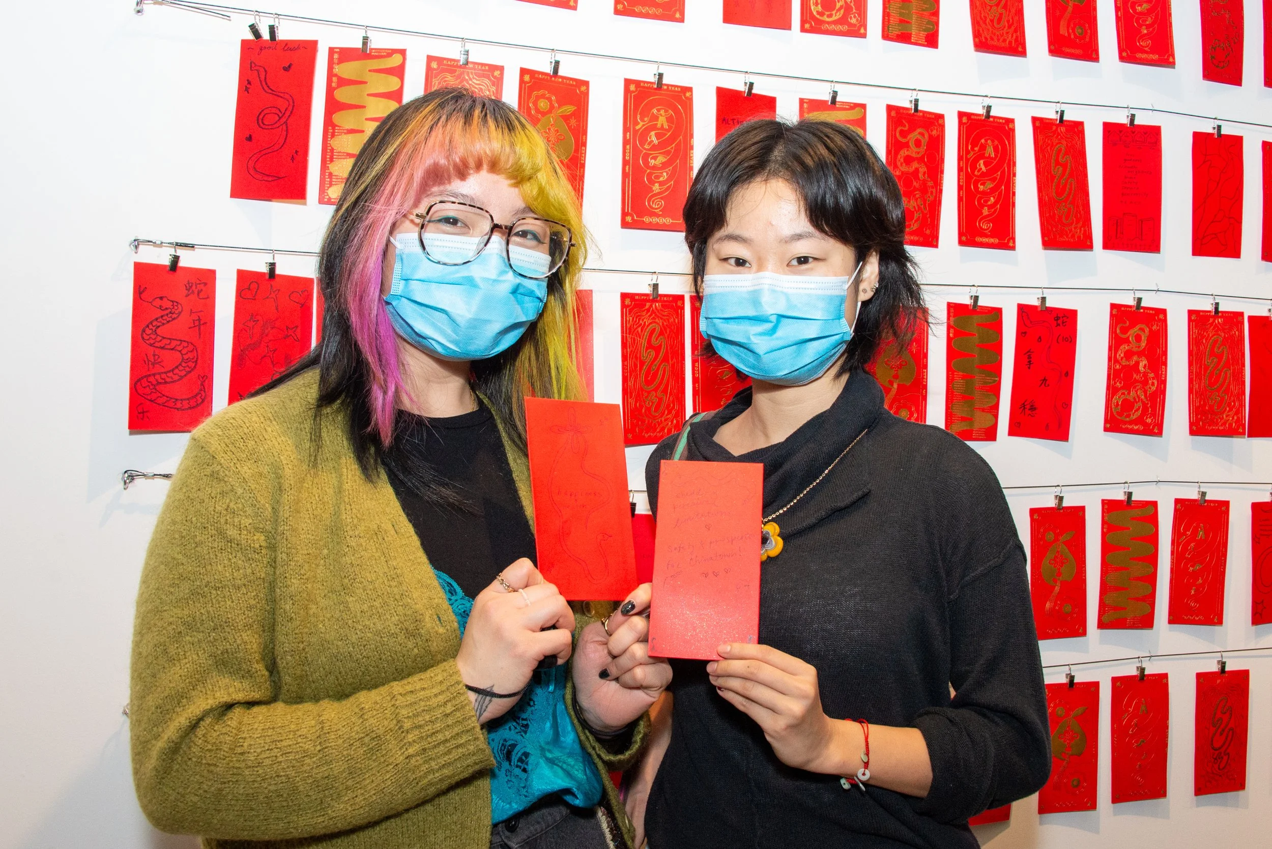 Two young women wearing face masks hold red envelopes in front of a display of red envelopes with gold designs, celebrating Chinese New Year.