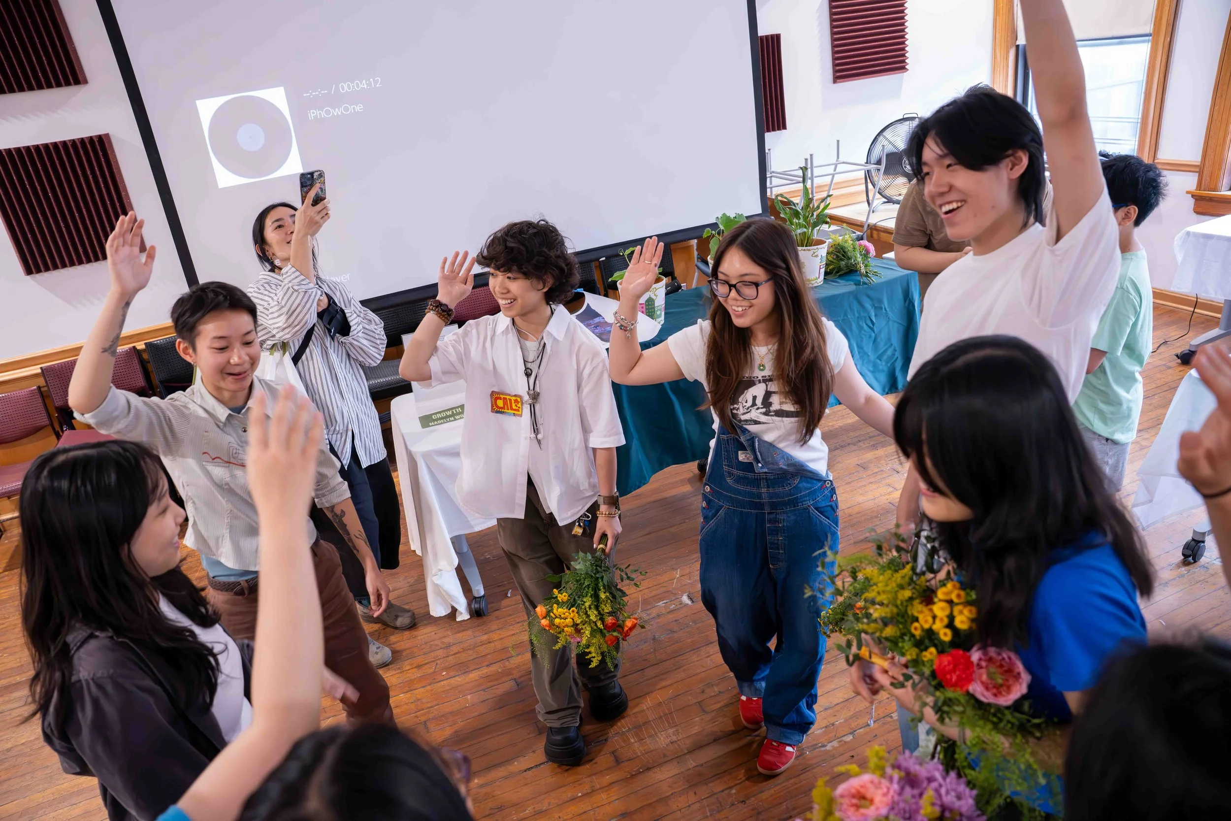 Group of people joyfully dancing and waving hands in a room with wooden floors, large windows, and potted plants, some holding bouquets of flowers.