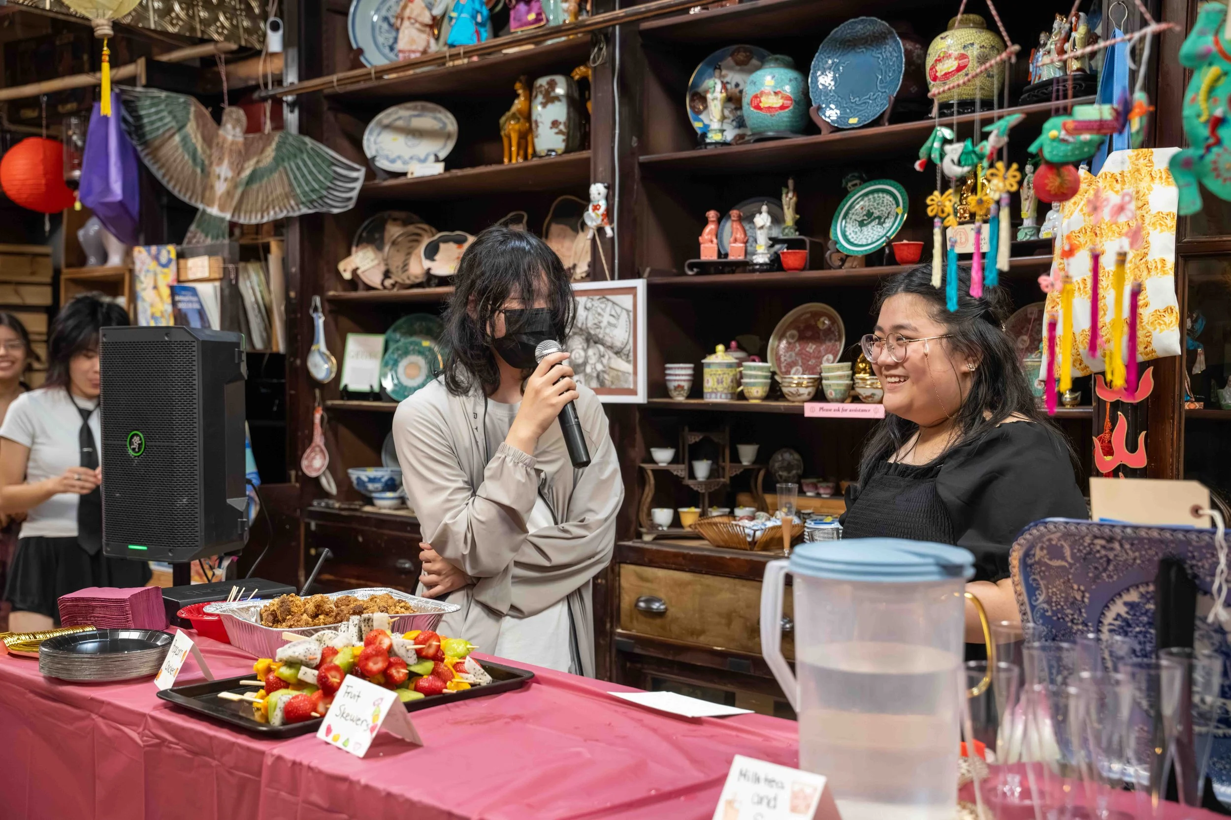 Women at an Asian-themed market or event, with one woman speaking into a microphone and another woman smiling nearby. The table in front has strawberries, fried food, and signs, with shelves in the background holding decorative Asian items.