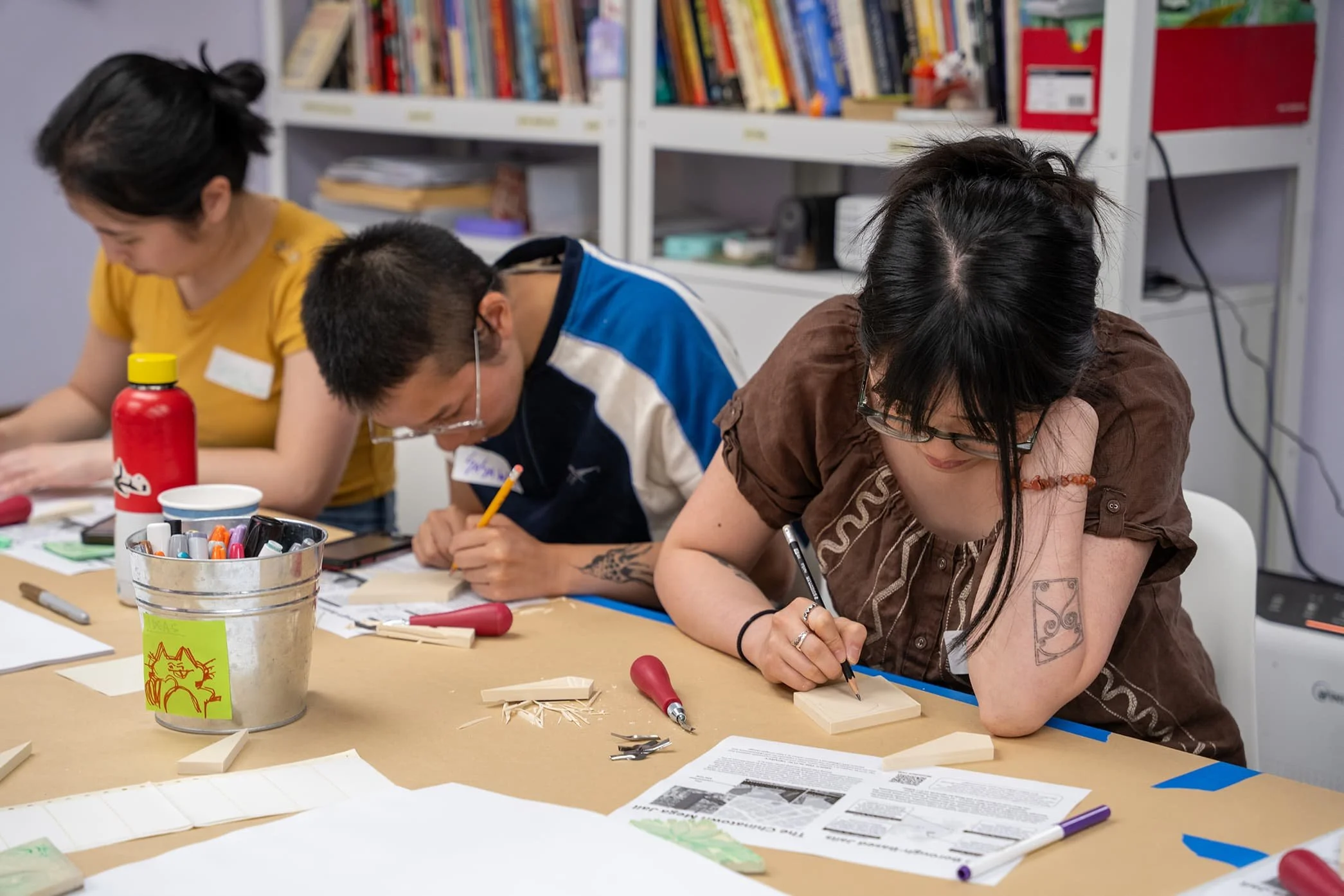 Three women and one man working on a woodworking project at a table, using pens and tools, with books and supplies in the background.