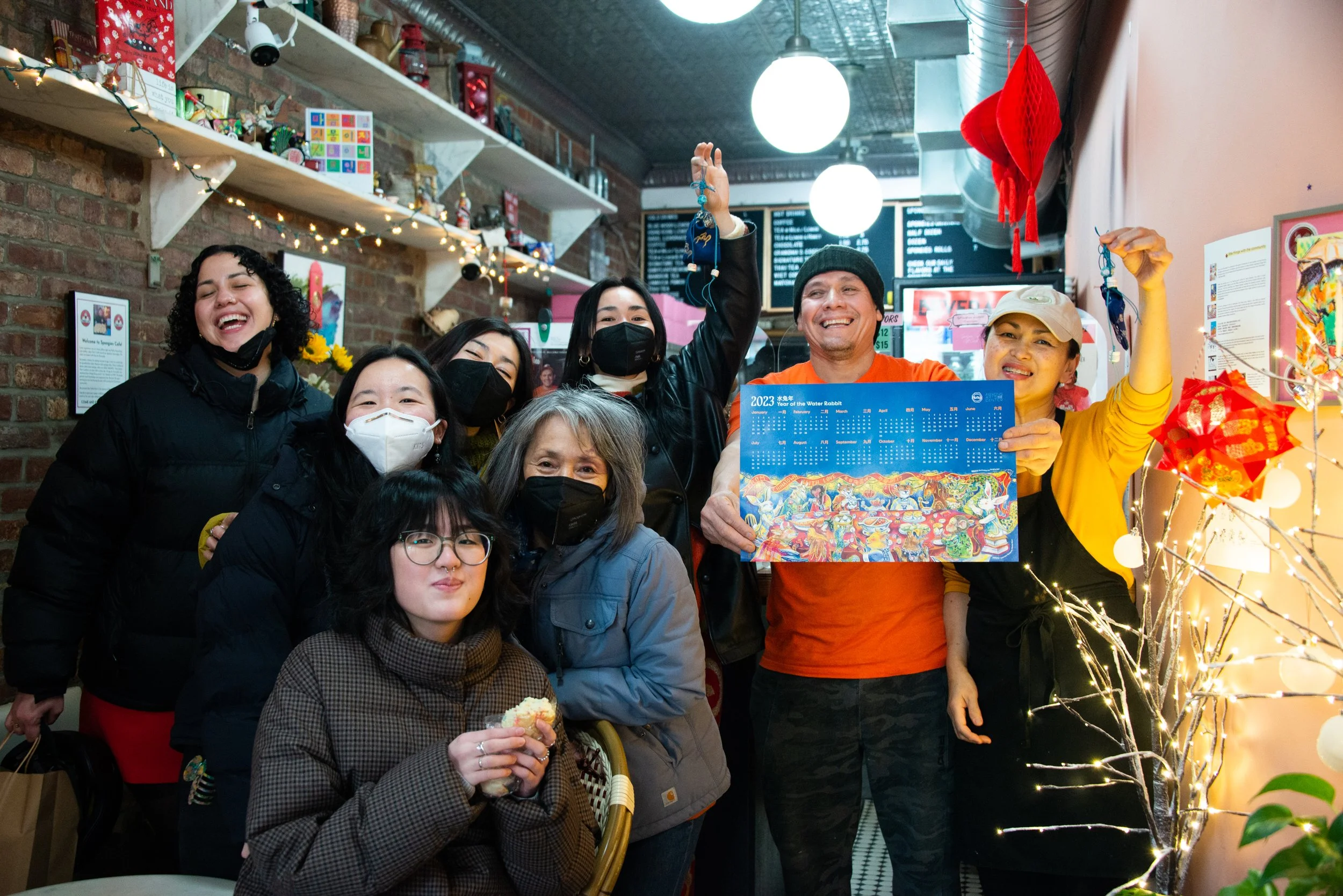 Group of people celebrating in a restaurant, holding a colorful 2023 calendar, smiling, some wearing masks, with festive decorations including string lights and red hanging ornaments.