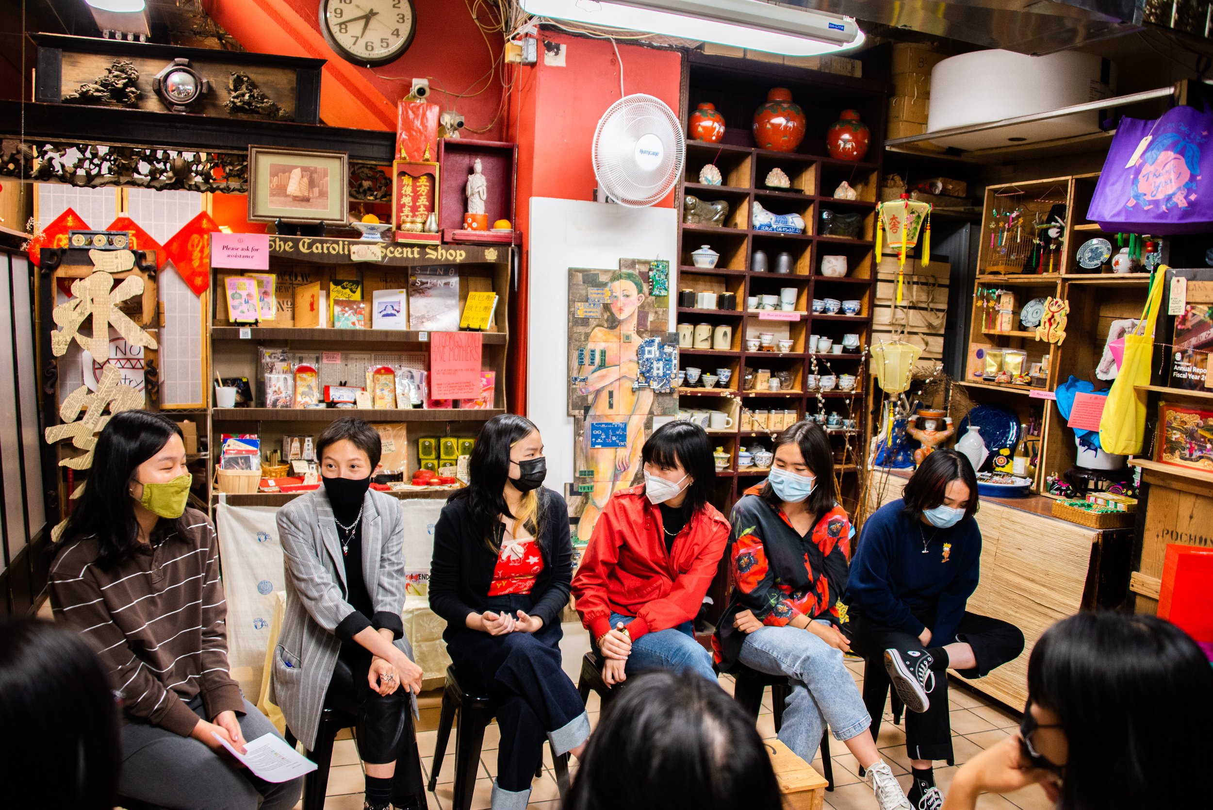 Six women sitting in a row inside a shop decorated with Asian art and pottery, all wearing face masks and engaging in conversation.