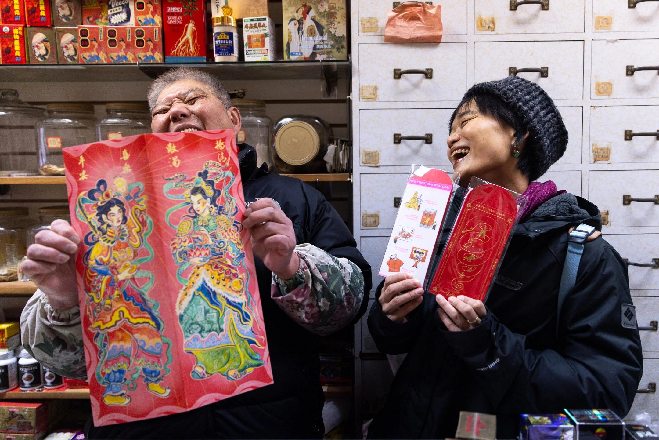 Two people in a store holding Chinese New Year decorations and laughing, with shelves of traditional items behind them.