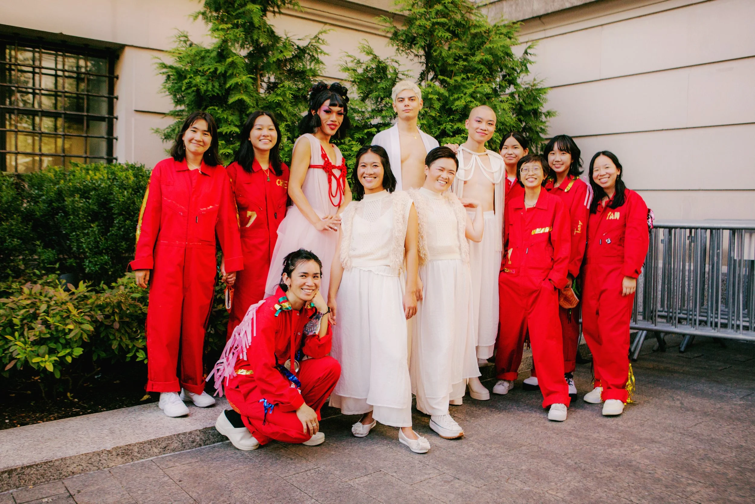 Group of people, including drag performers and students in red uniforms, posing outdoors in front of green bushes and a building.