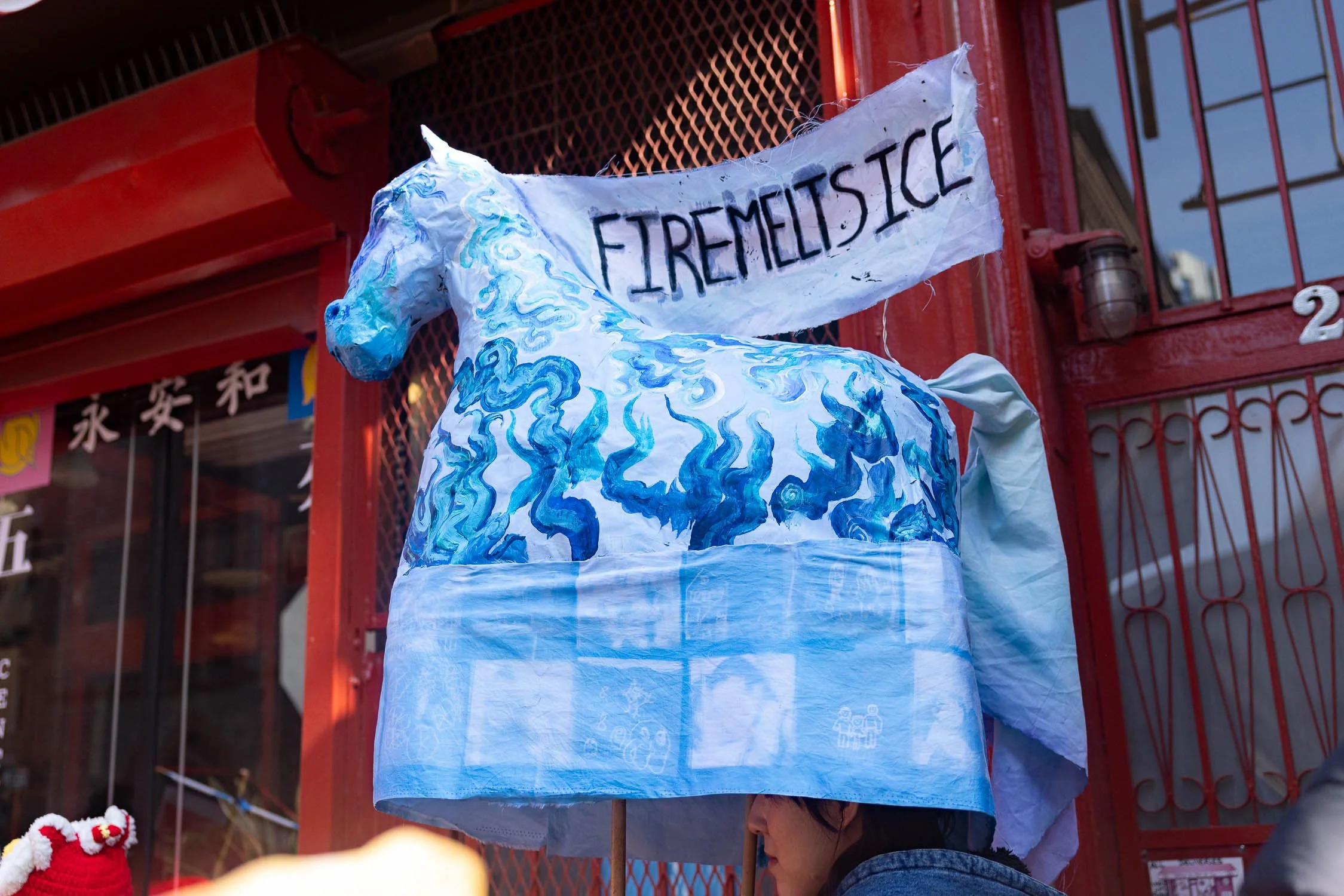 Fourth fire horse in Chinatown. The horse is blue and covered in painted blue flames. The horse's fabric mane reads "fire melts ice." The horse has a cyanotype quilt draped at the bottom made of drawings of family from community members