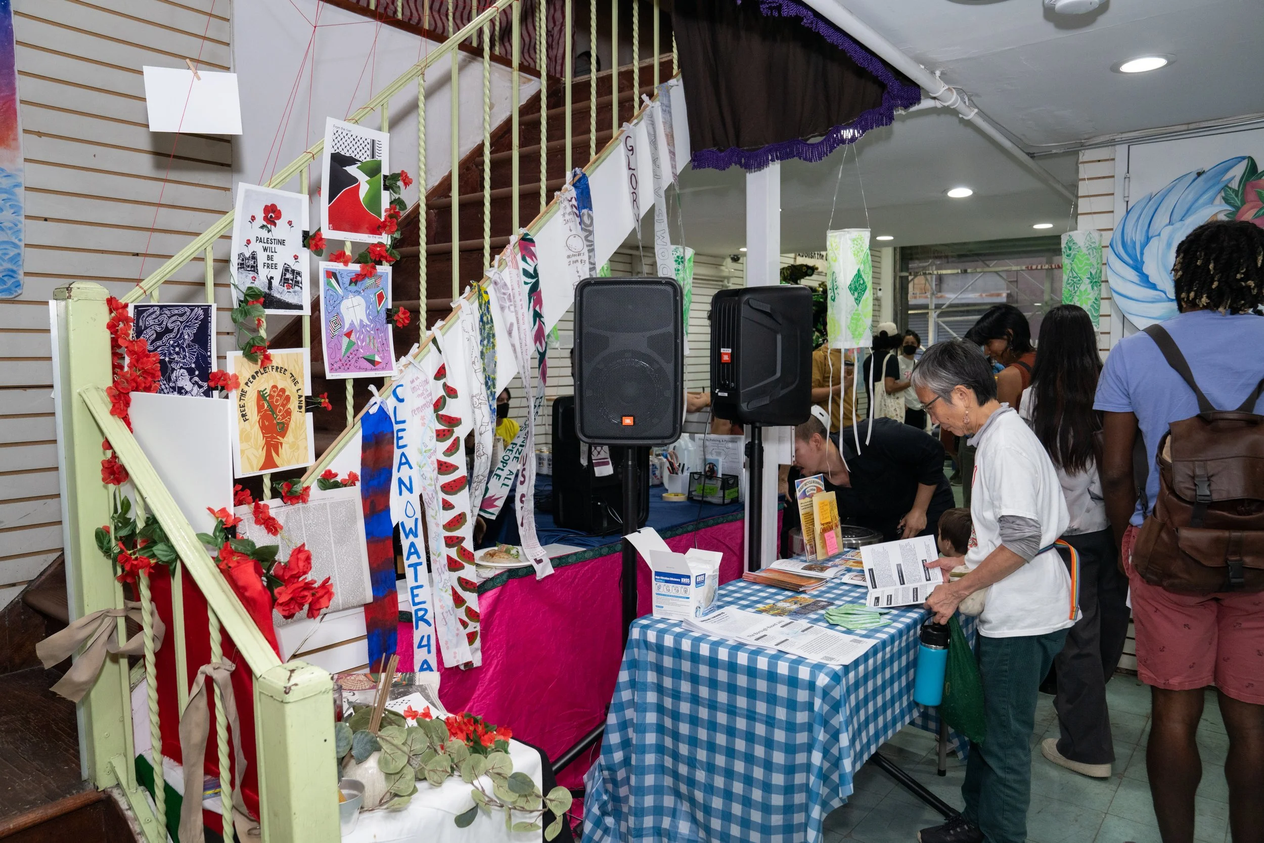 A booth at an indoor event features colorful banners and artwork hanging from the staircase railing, with a table covered in a blue checkered cloth displaying brochures and informational material. Several people are browsing and engaging with the displays, and there are large speakers placed near the booth.