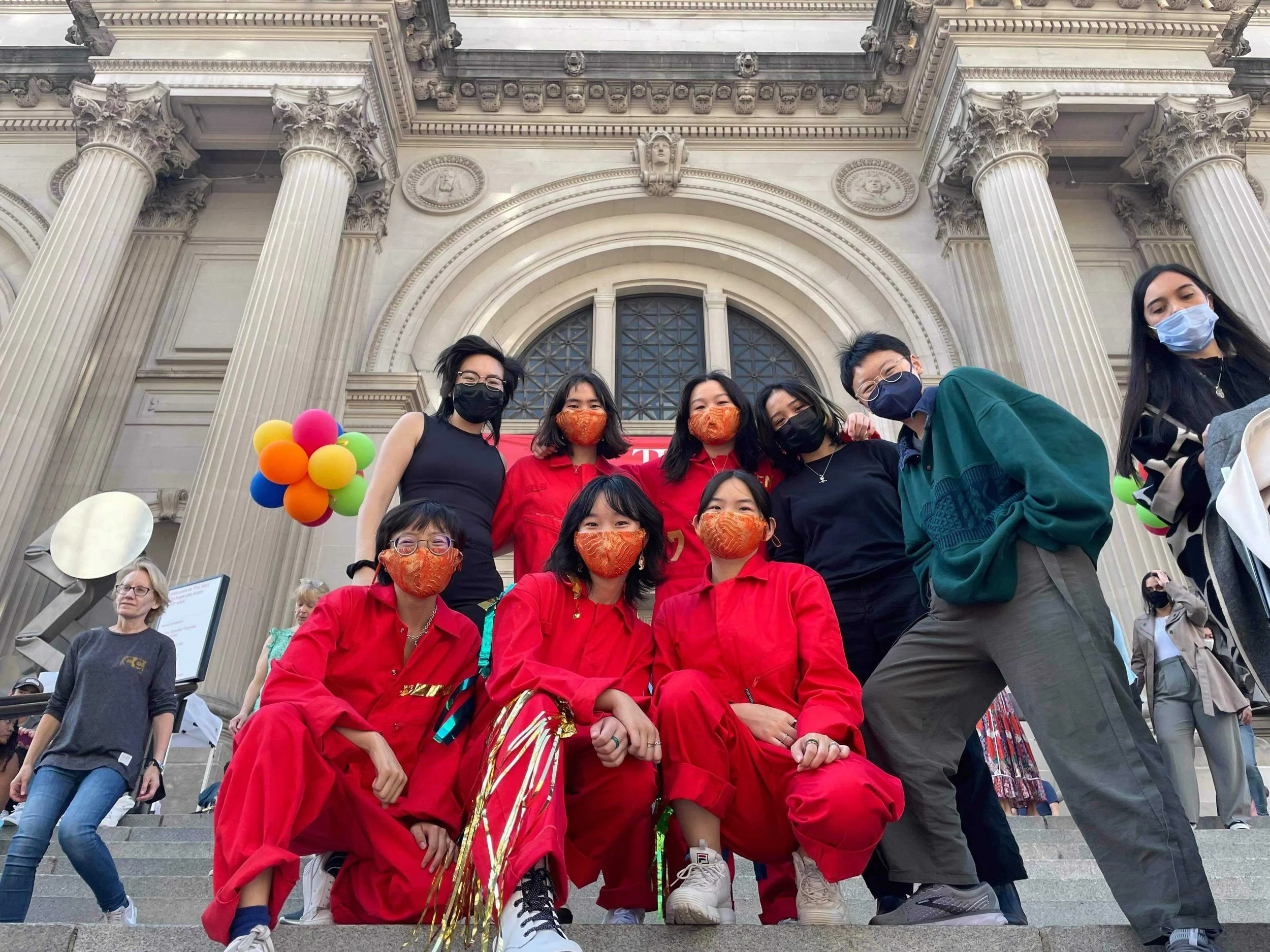 A group of children and adults wearing masks poses on the steps in front of a grand building with columns, some of the children are dressed in red outfits.