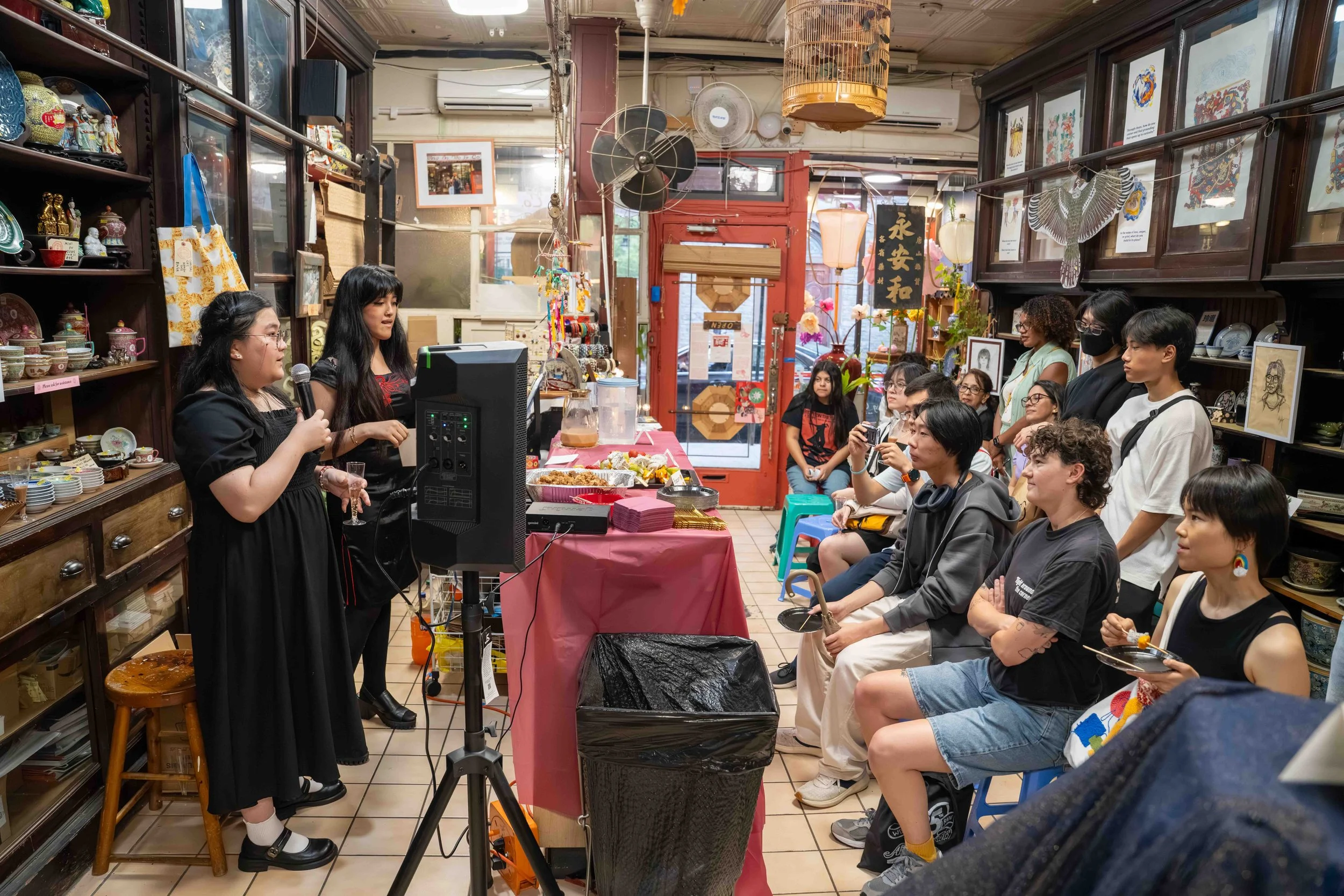 Two women are giving a presentation with a microphone to an audience in a decorated shop with shelves of ceramics and artwork.