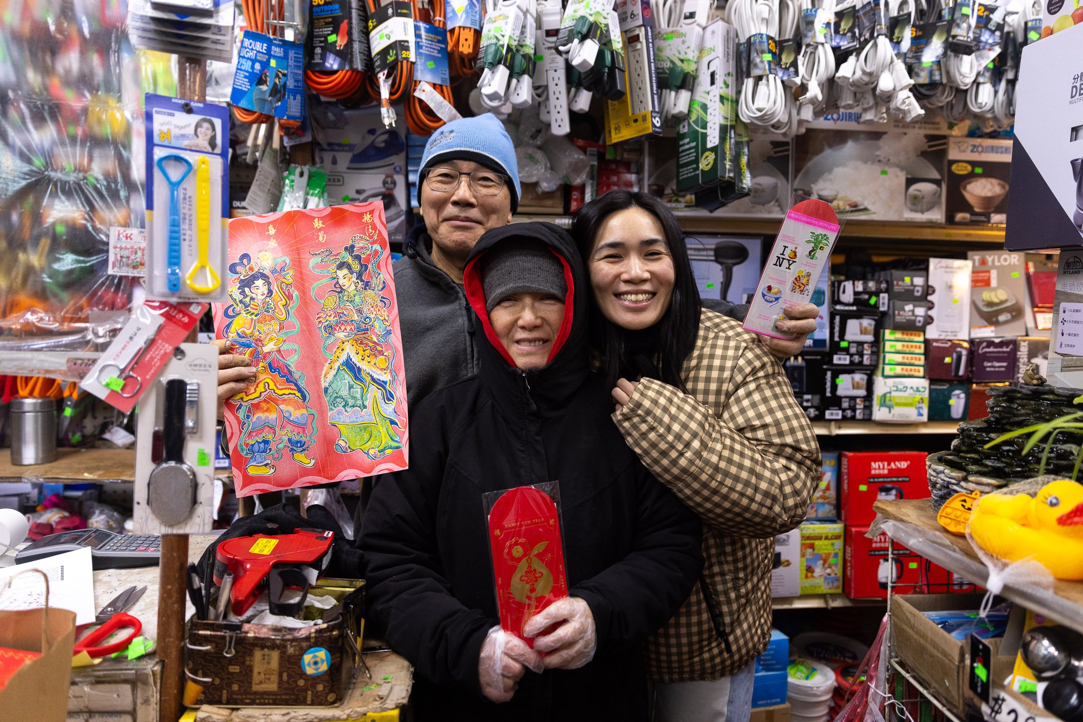 Three people inside a store, two women and a man, celebrating Lunar New Year. They are holding red and colorful decorations, with shelves of assorted household items and gifts in the background.