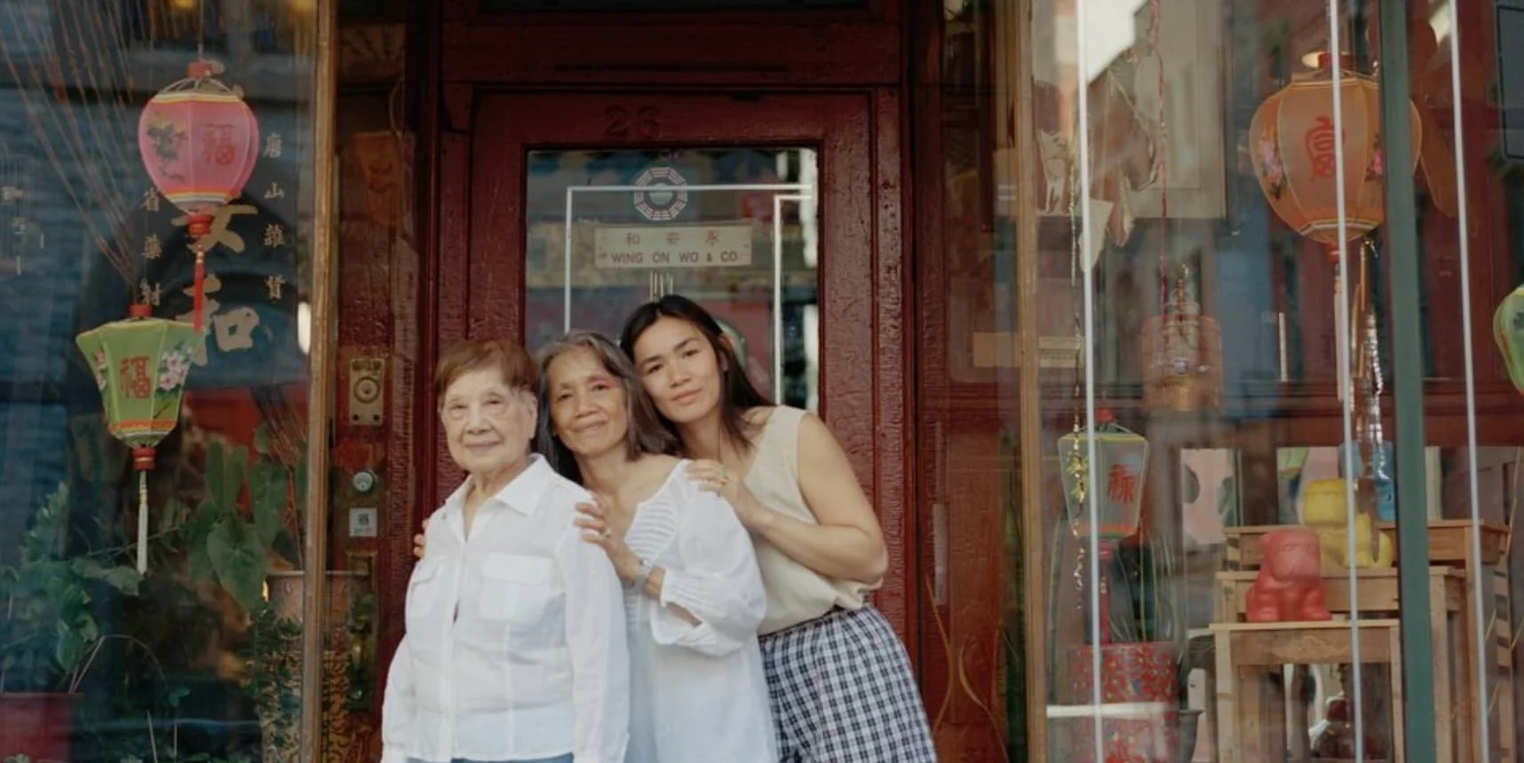Three women standing together in front of a store with Chinese decorations, smiling at the camera, with lanterns and ornaments displayed in the window.