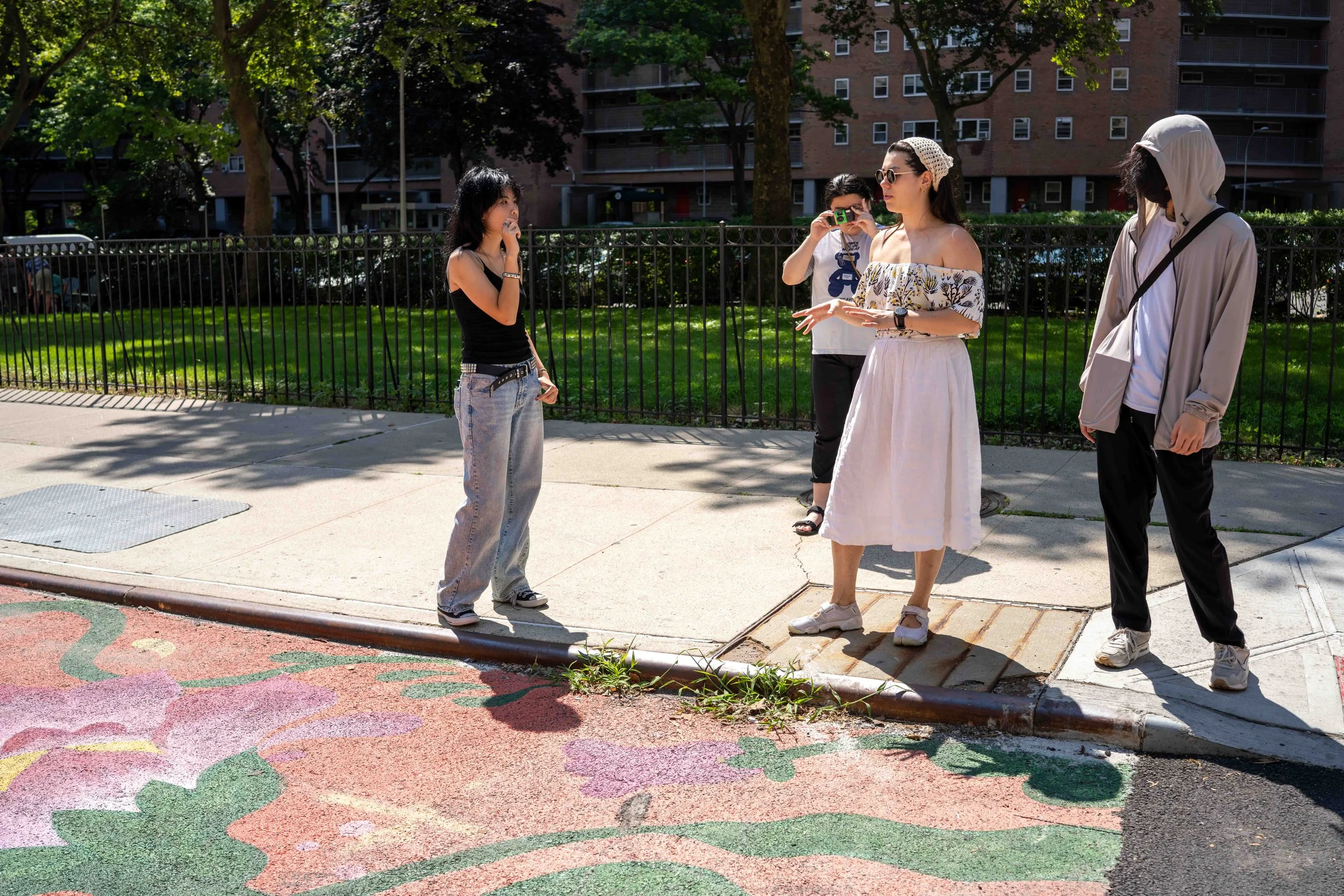 Four young people standing on a sidewalk, with a woman in a white skirt and sunglasses speaking while the others listen; one person is taking a photo, another wears a gray hoodie, and the fourth is in a black tank top and jeans. They are outdoors, with trees, a grassy area, and apartment buildings in the background.