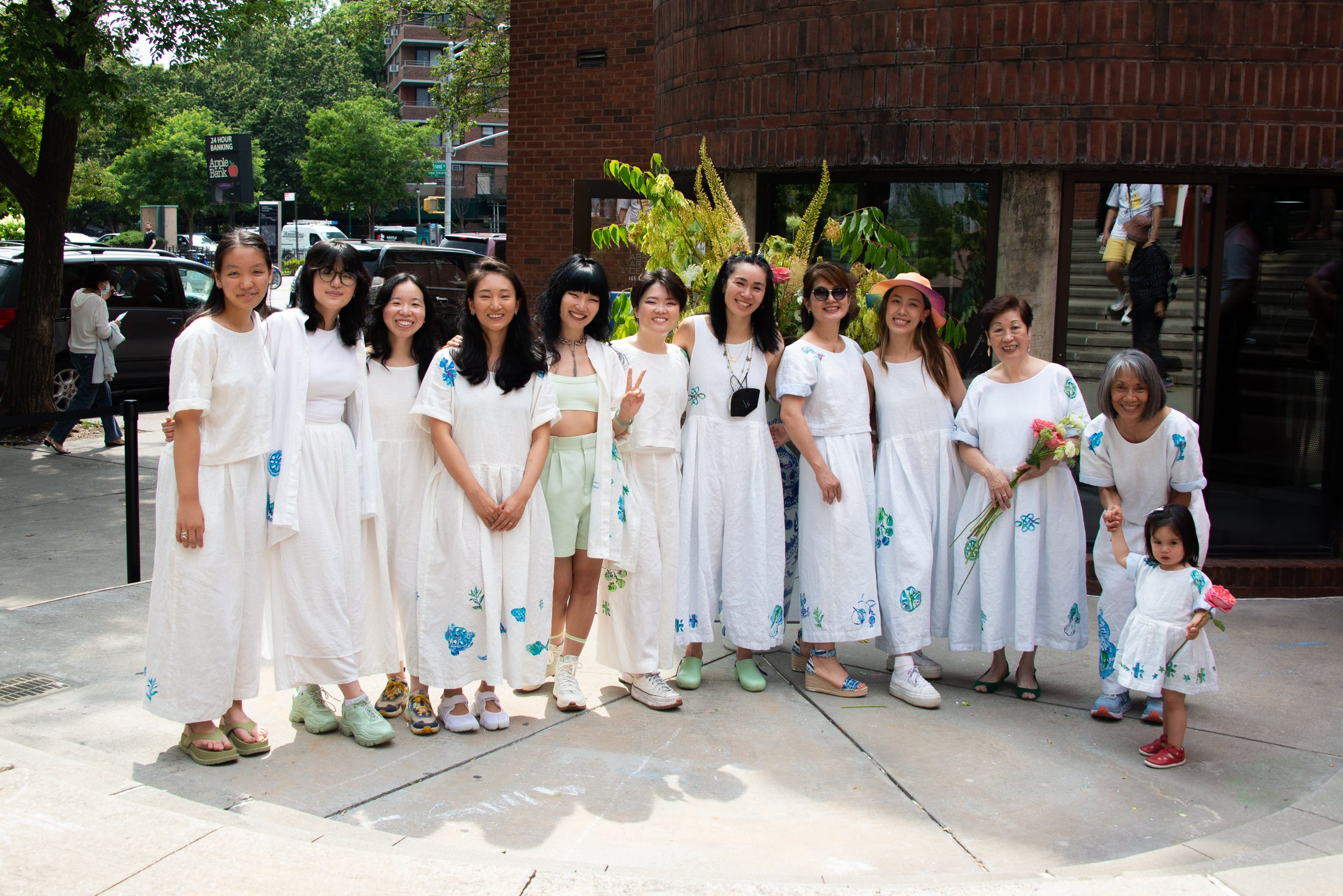 A group of people dressed in white clothes standing outdoors in front of a building with lush greenery behind them. They are smiling, some holding flowers, and posing for a photo on a sunny day.