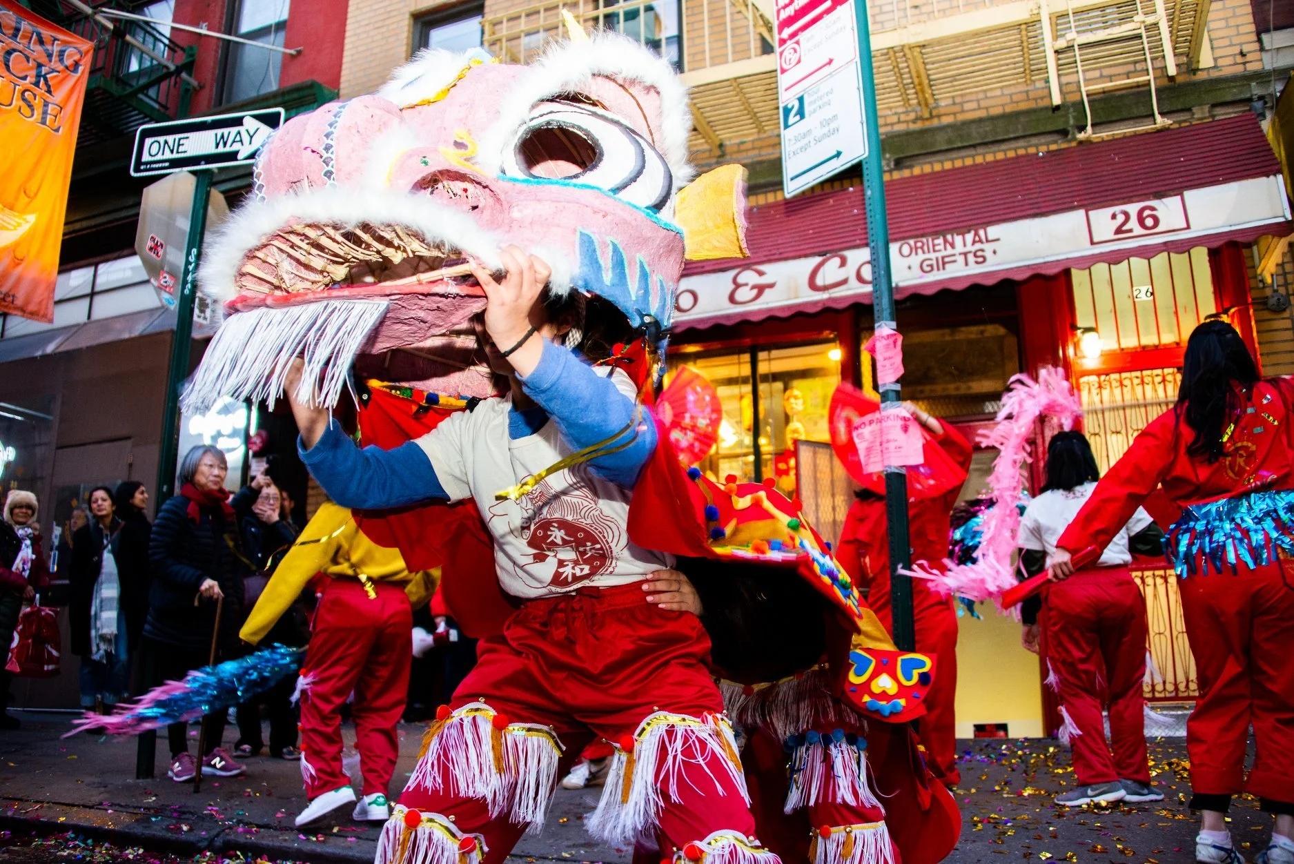 People celebrating a Chinese New Year parade, with a performer wearing a lion dance costume and several others holding pink feathered props on a city street.