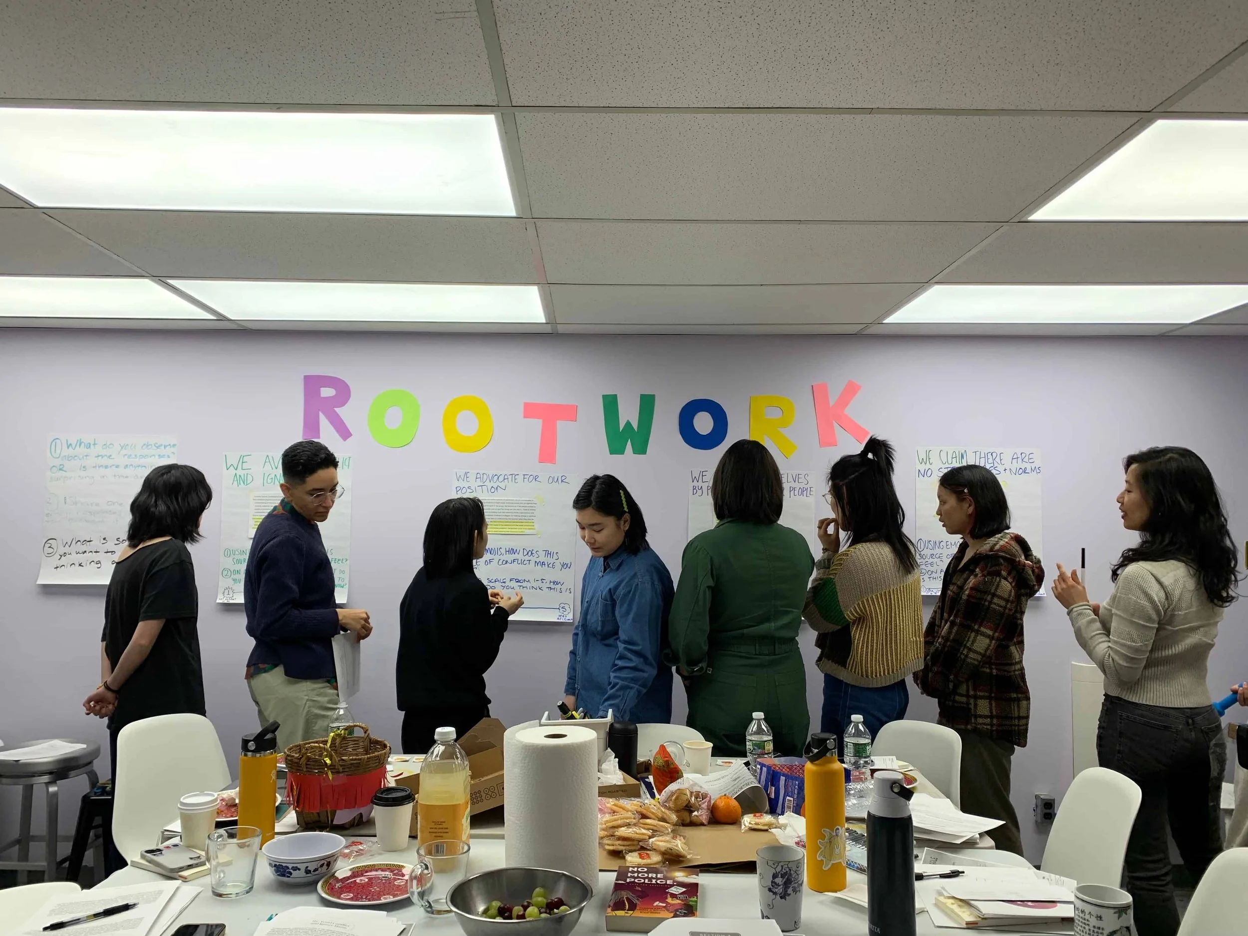 A group of women standing in line at a workshop, with a wall behind them decorated with large, colorful letters spelling 'ROOTWORK'. A table in front has snacks, drinks, and papers.